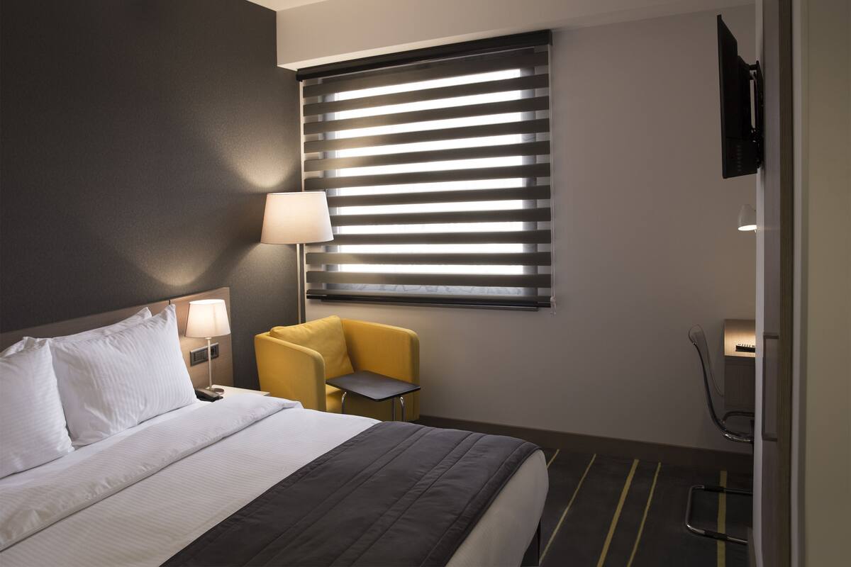 Une chambre d’hôtel avec un lit, une chaise, une lampe, un téléviseur et une fenêtre avec des stores.