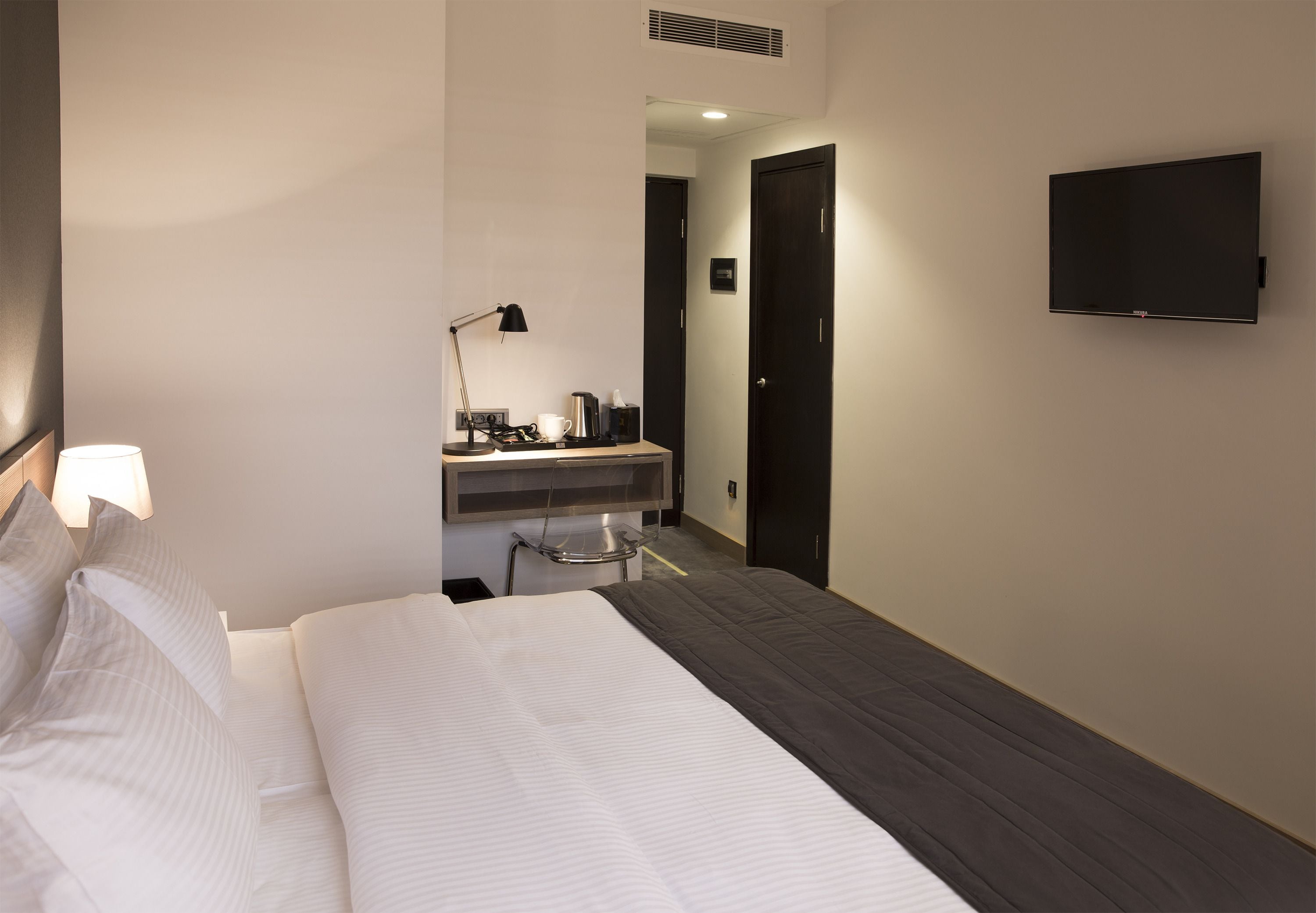 A hotel room with a bed, bedside lamp, TV, and a small kitchenette area.
