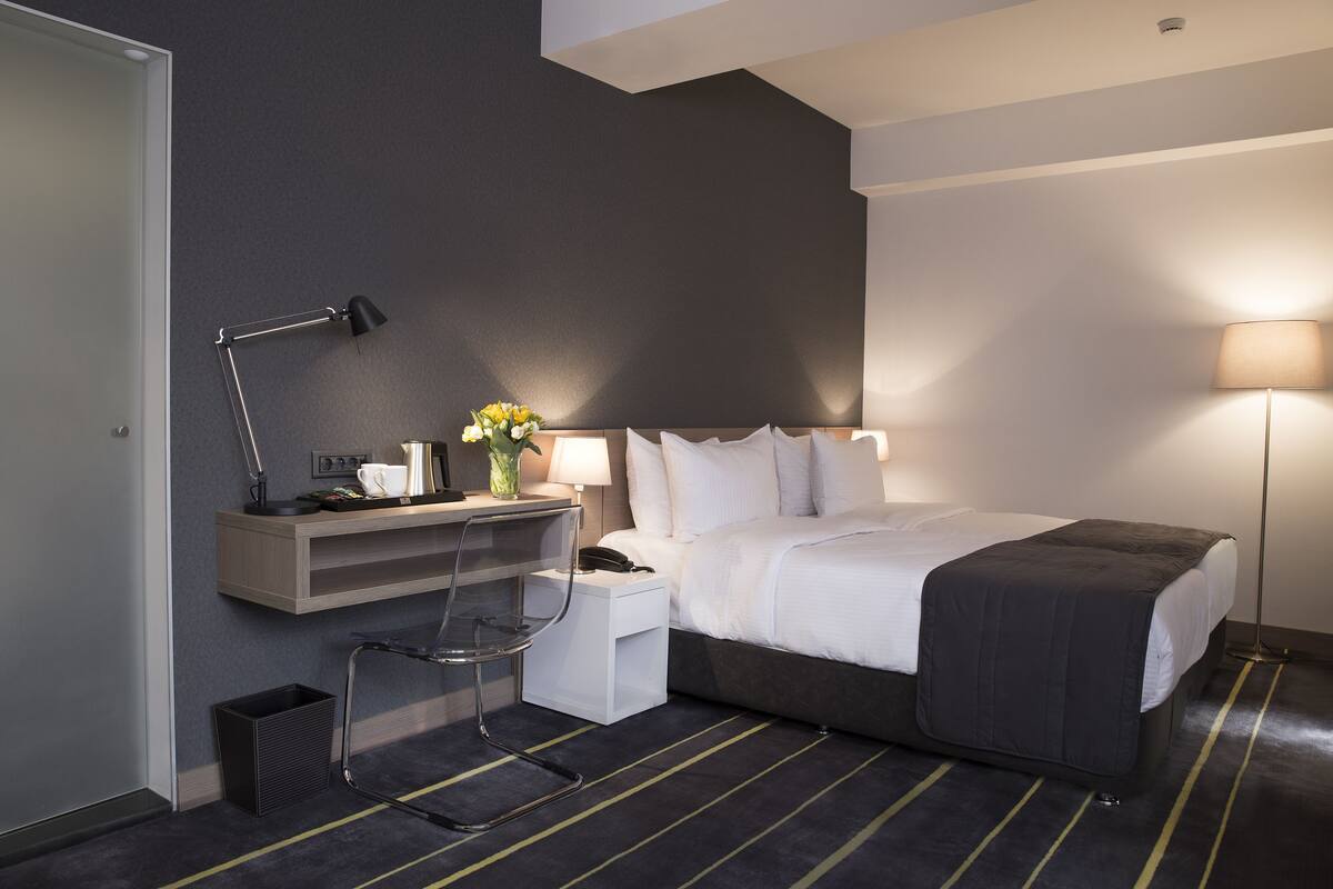 Une chambre d’hôtel moderne équipée d’un lit, d’une table de chevet, d’un bureau, d’une chaise, d’une lampe et d’une petite poubelle.