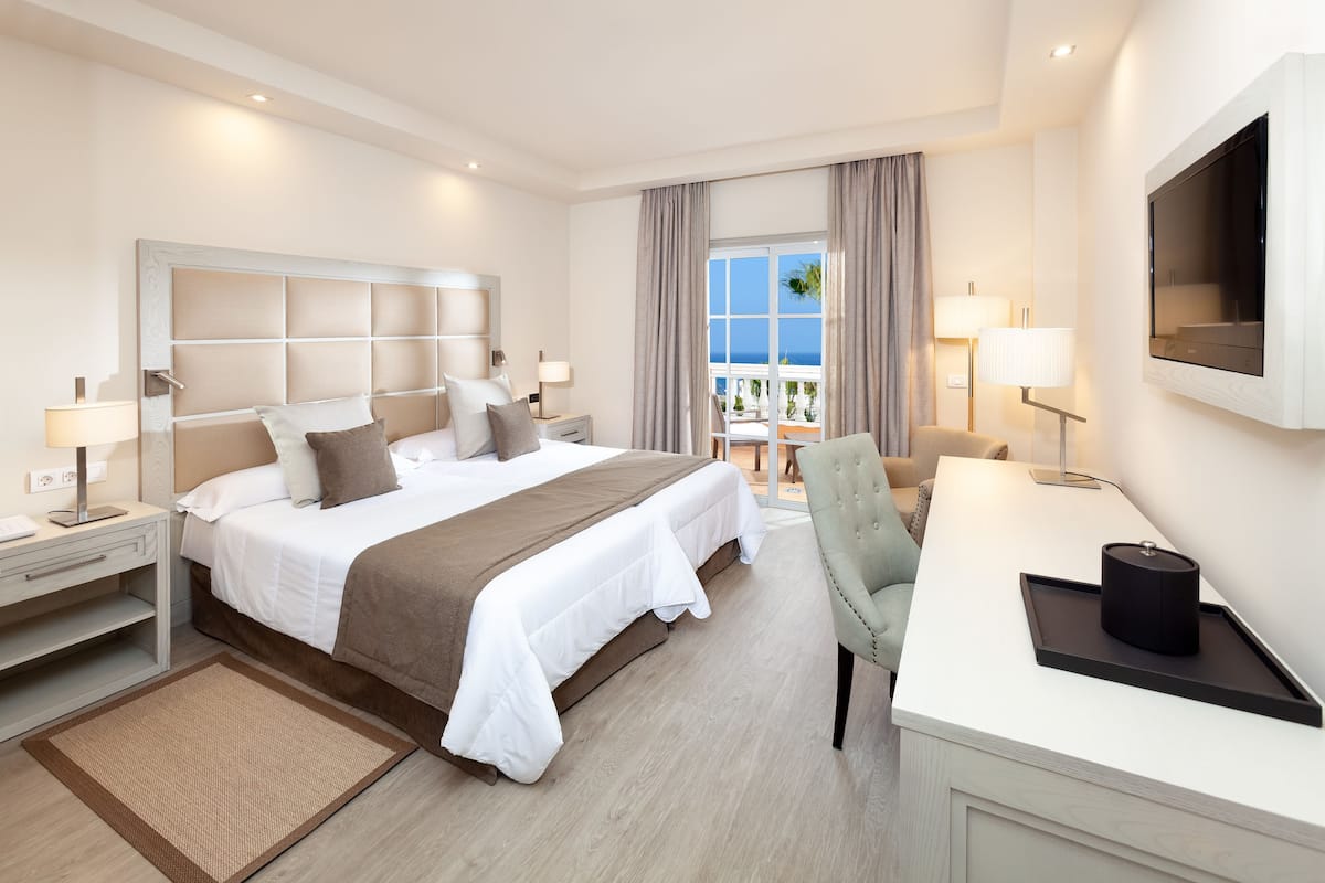 Habitación de hotel con una cama grande, un escritorio con silla, televisor y vista al mar desde la ventana.