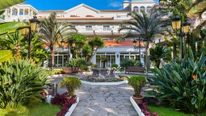 A courtyard with a fountain, palm trees, and a building with a tiled roof. - Hotel Riu Garoé (Puerto de la Cruz)
