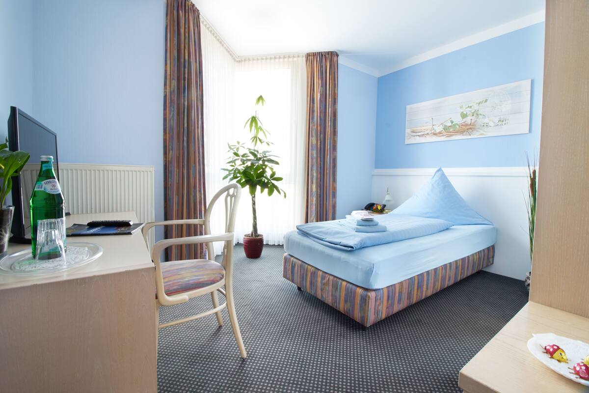 Een hotelkamer met een bed, een bureau met stoel, een televisie, een potplant en een raam met gordijnen.