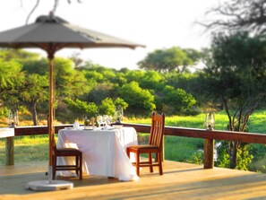Un área de comedor al aire libre con una mesa dispuesta para dos personas, rodeada por una terraza de madera y una barandilla.