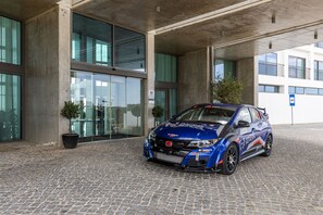 Un'auto da corsa blu parcheggiata di fronte a un edificio con ampie vetrate e una pianta in vaso.