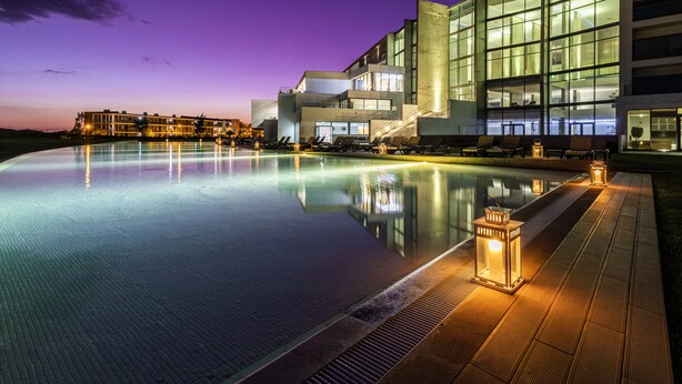 Een modern hotel met een groot overloopzwembad, terras en een goed verlichte gevel.