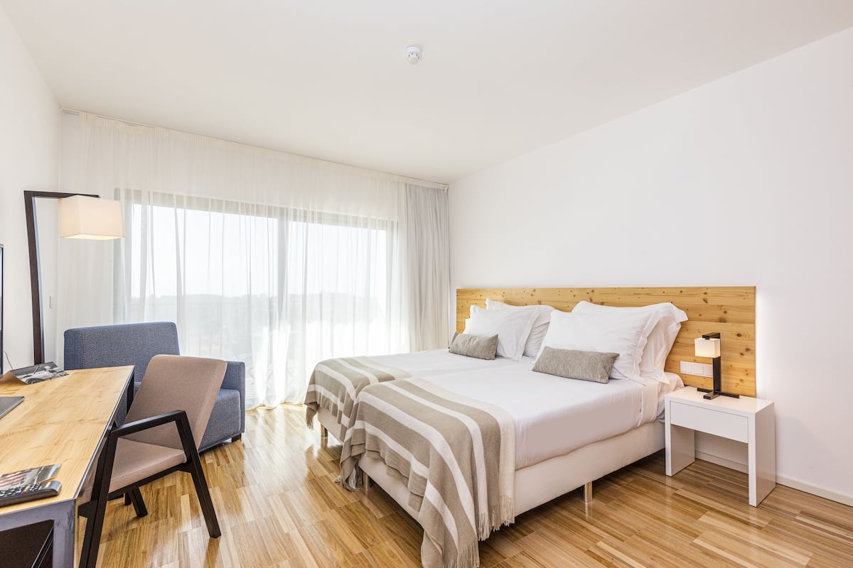 Een hotelkamer met houten vloer, een bed met witte beddengoed, een bureau met stoel, een nachtkastje met lamp en een groot raam met dunne gordijnen.