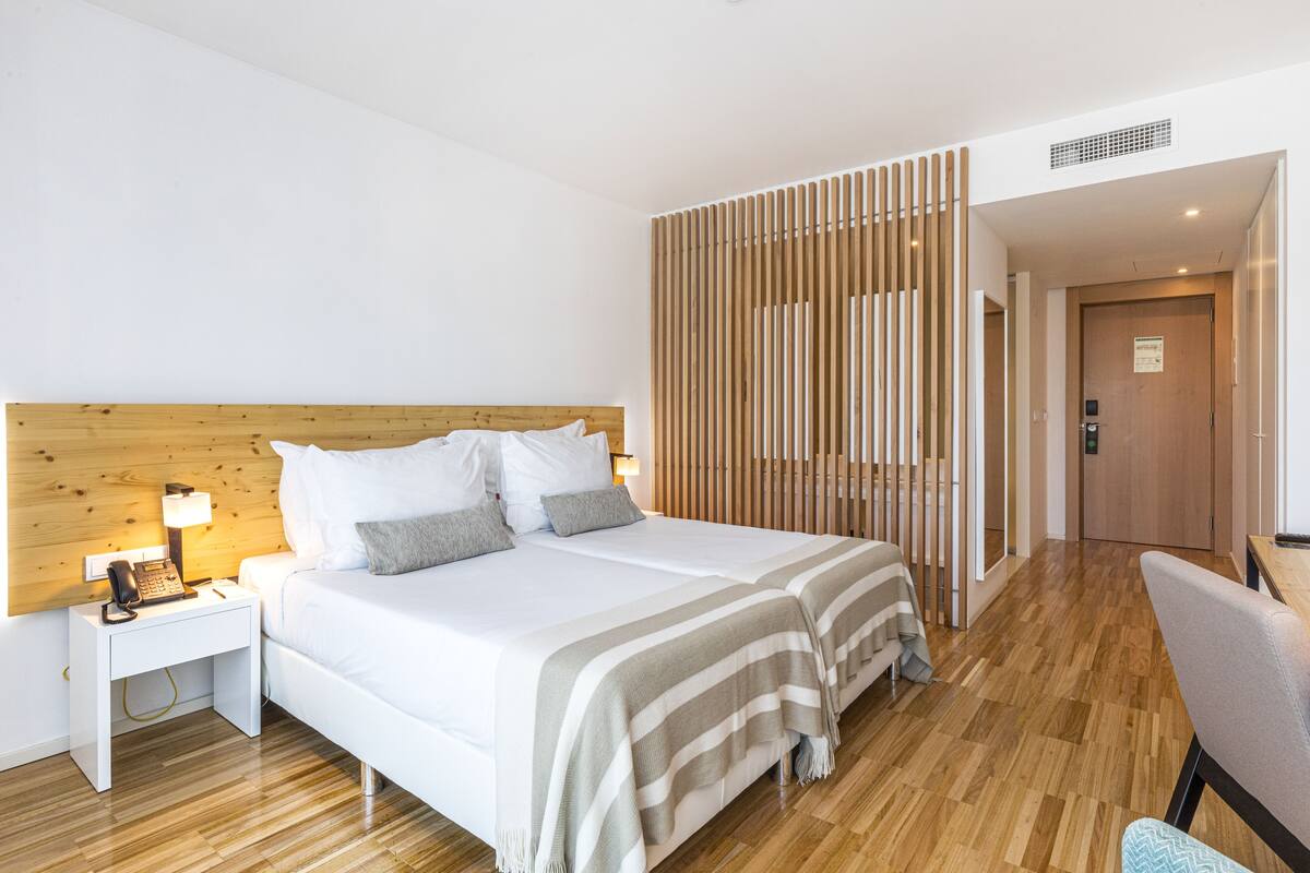 Een hotelkamer met een houten hoofdeinde, een bed met witte lakens, een nachtkastje met een lamp en een houten vloer.