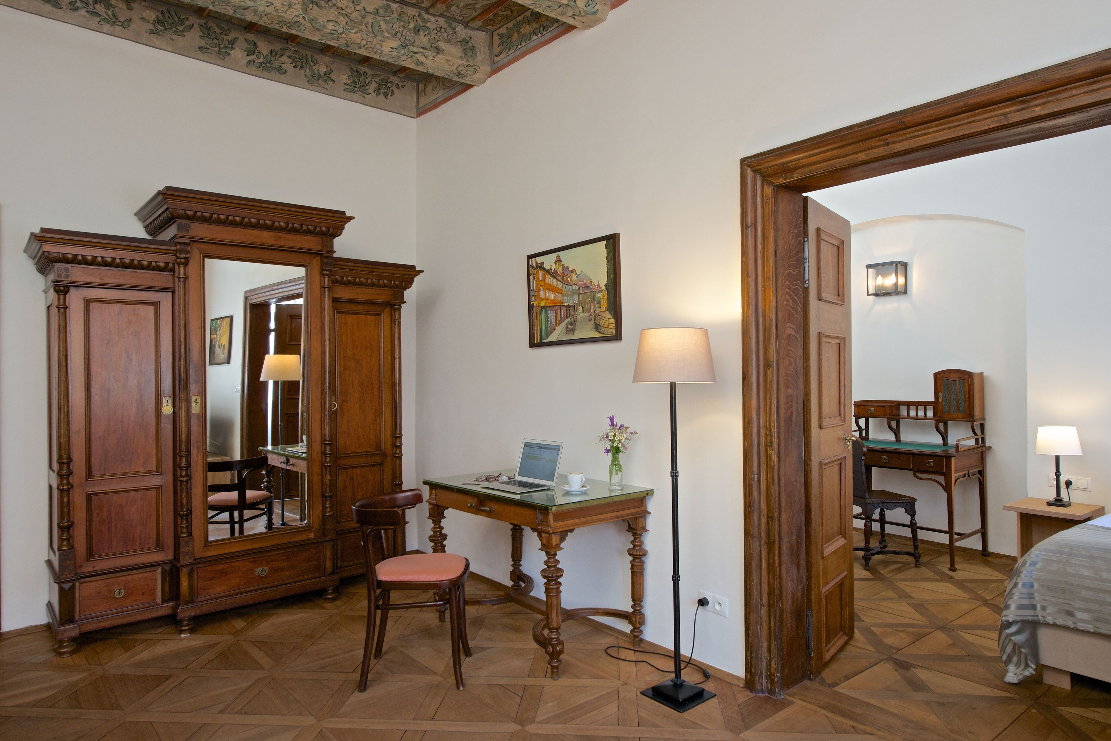 Una habitación con un escritorio de madera, una silla y un espejo grande. Hay una lámpara, un cuadro en la pared y una cama en la esquina.