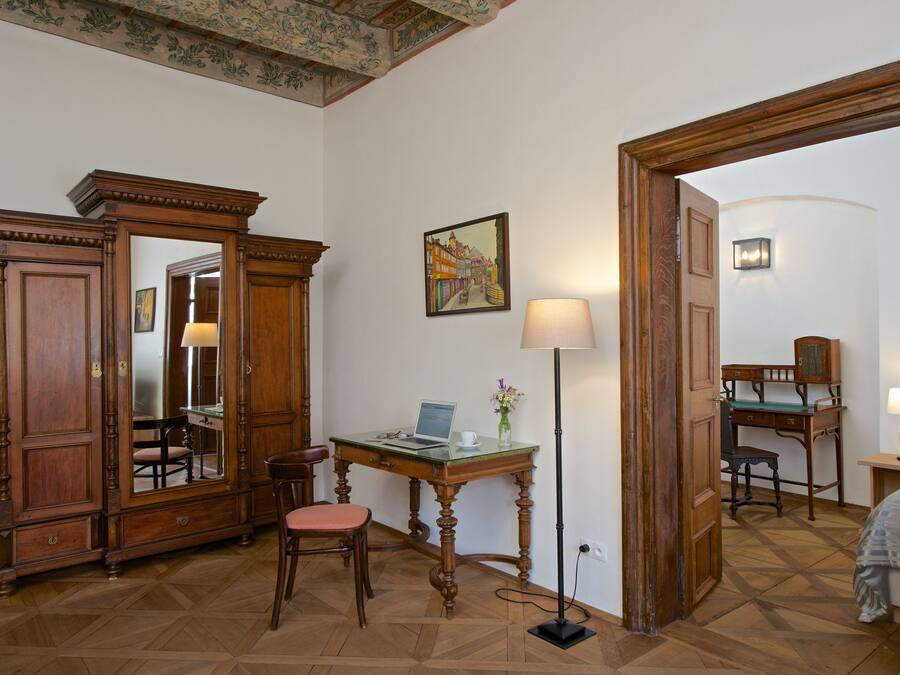 Una habitación con un escritorio de madera, una silla y un espejo grande. Hay una lámpara, un cuadro en la pared y una cama en la esquina.