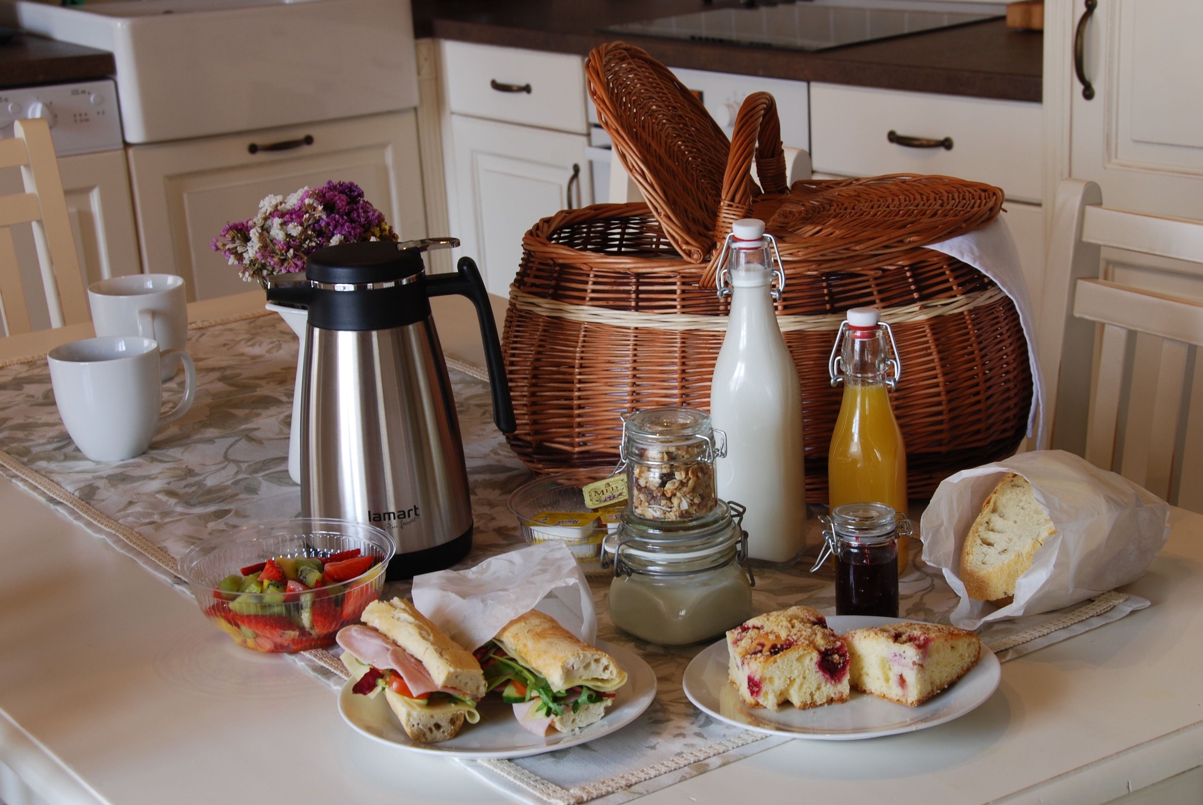 A breakfast table with sandwiches, a fruit bowl, a coffee maker, a bottle of milk, a basket, and a jug of juice.
