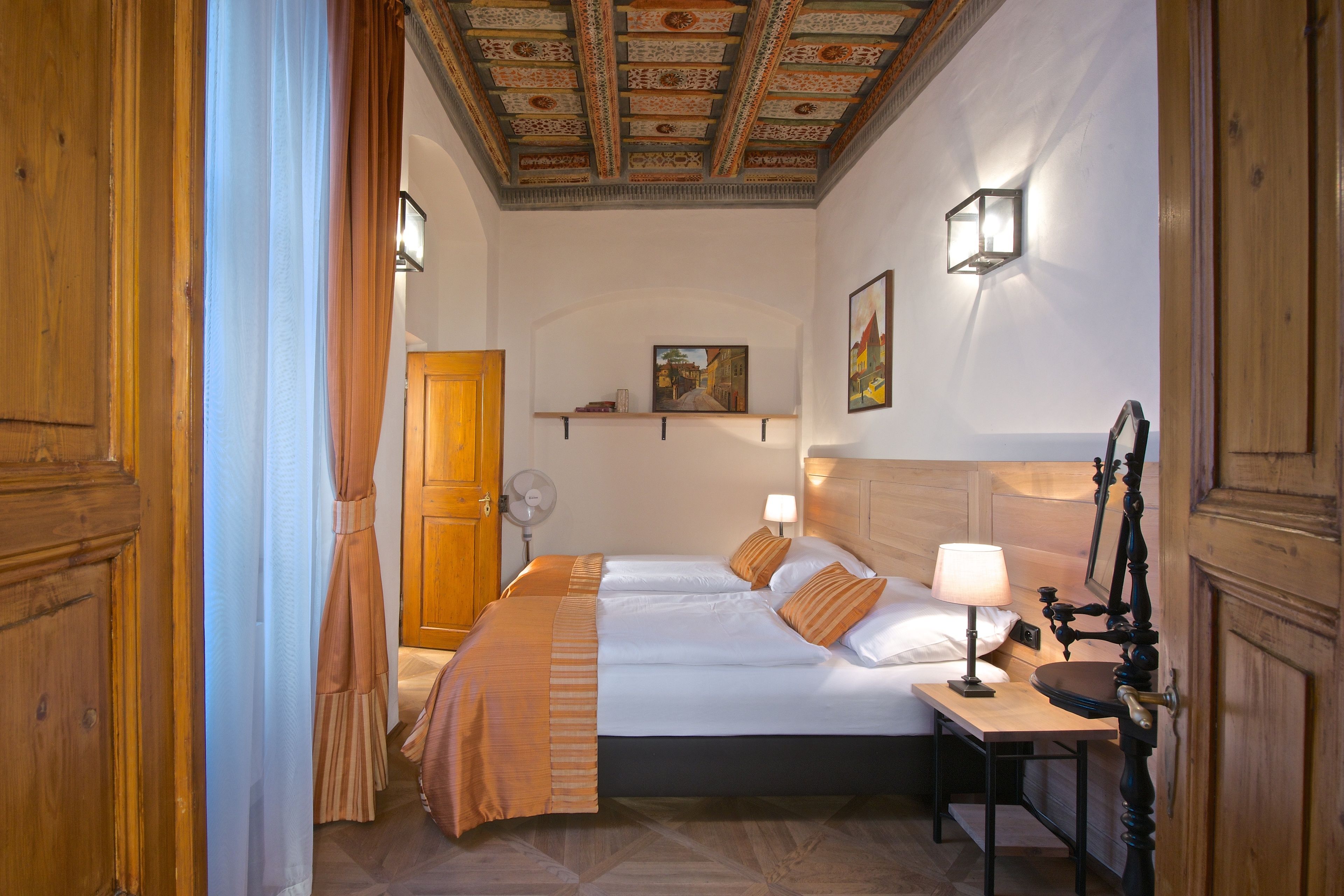 A hotel room with a wooden ceiling, two beds with matching headboards, a bedside table with a lamp, and a wall-mounted shelf with a painting.