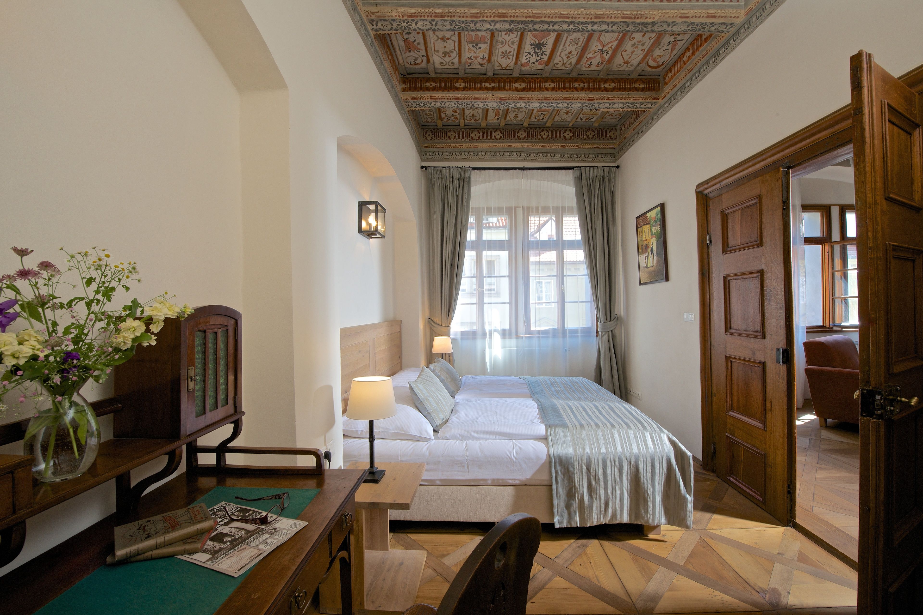 A hotel room with a bed, a desk, a chair, a vase with flowers, a lamp, and a wooden door.