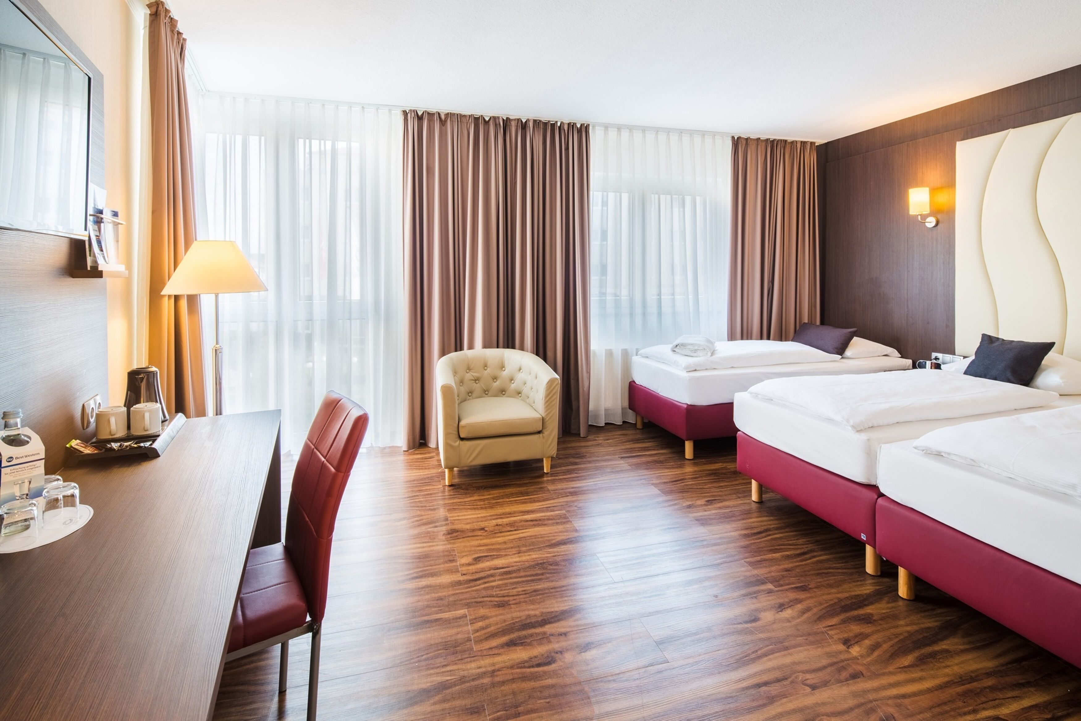 Ein Hotelzimmer mit Holzboden, einem Schreibtisch mit Lampe, einem Sessel, zwei Betten und einem großen Fenster mit Vorhängen.