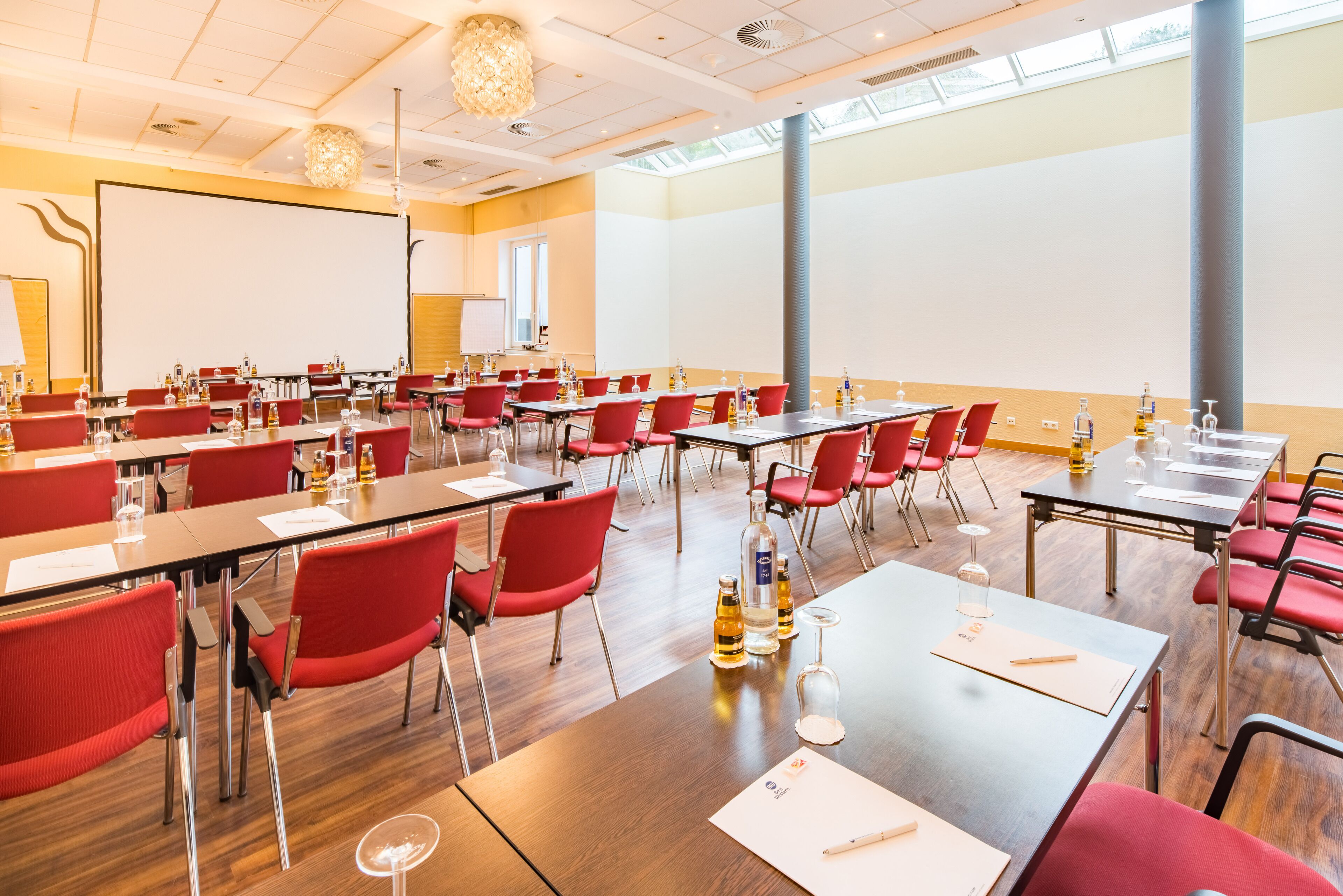 A conference room with red chairs, wooden tables, and a large screen.
