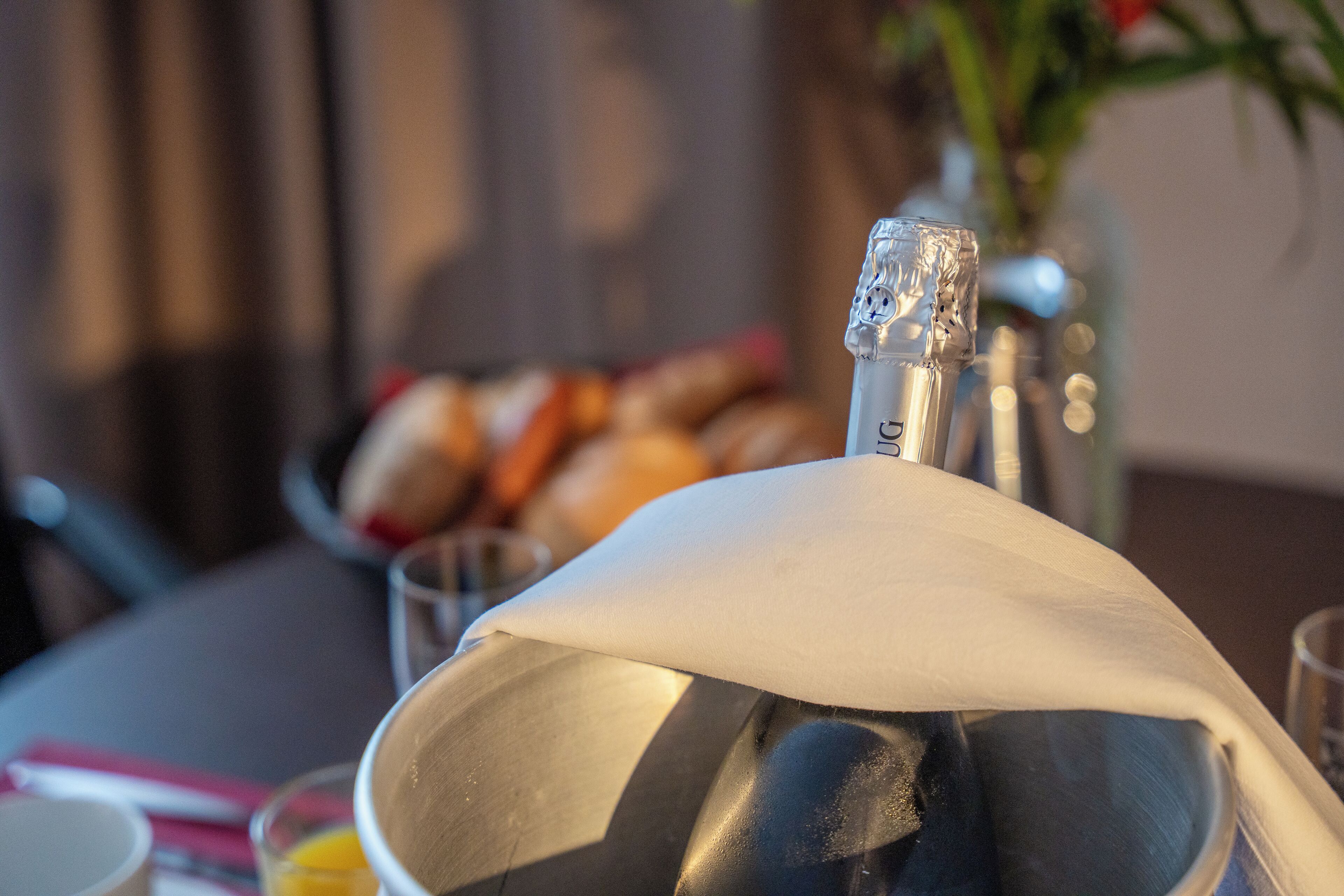 Metallinen esine, jonka päällä on valkoinen kansi, mahdollisesti jokin säiliö tai laite, seisoo pöydällä. Tausta on epätarkka, ja siinä näkyy kulho, jossa on leipää.