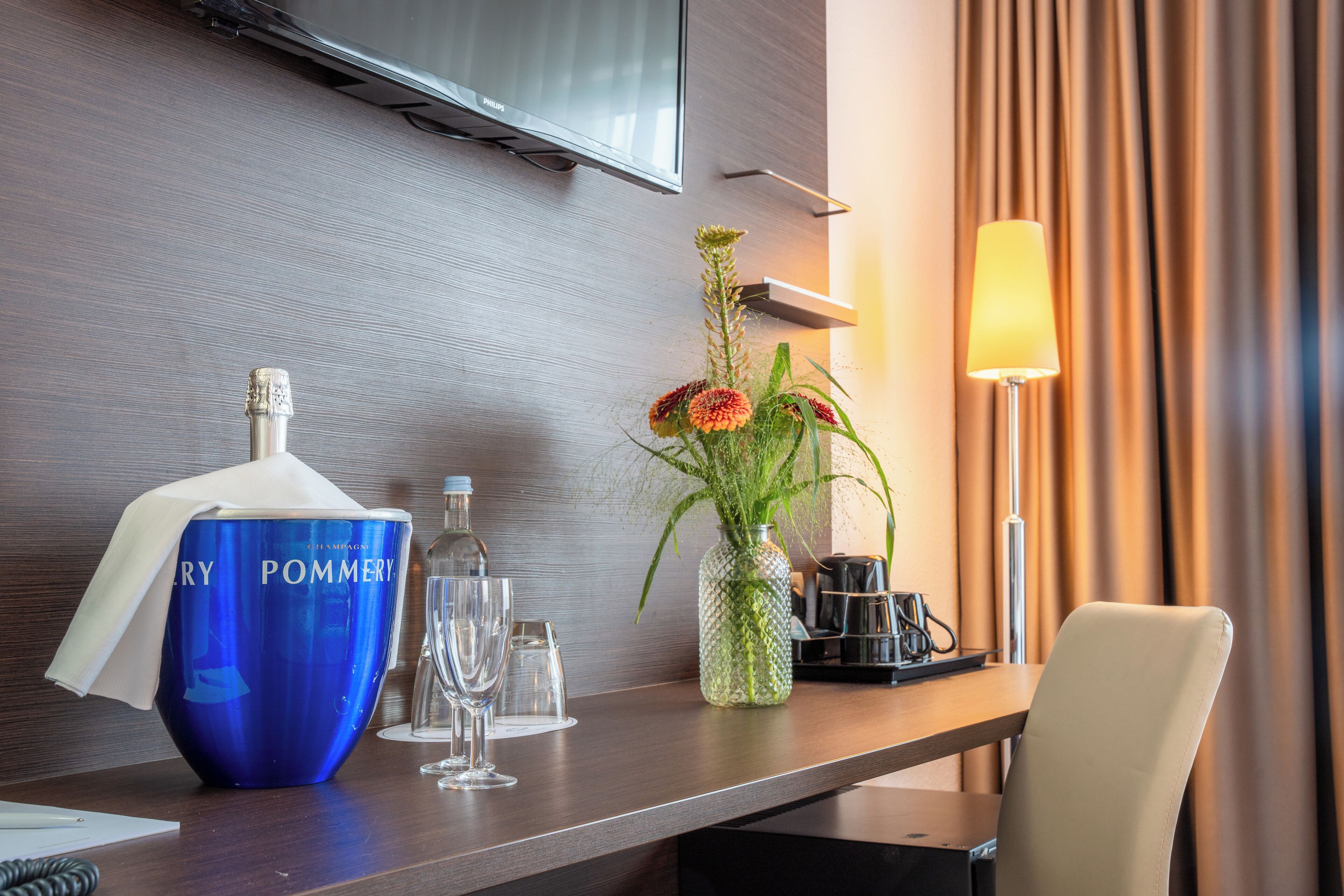 Hotelkamer met een houten bureau, een blauwe fles met de tekst 'POMMERY', een vaas met bloemen, een lamp en een aan de muur bevestigde televisie.