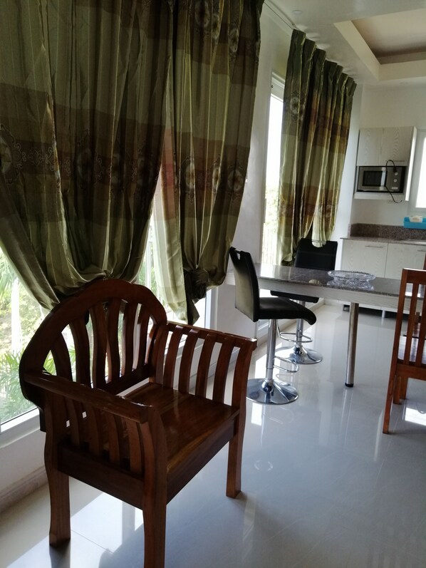 A modern kitchen with white cabinets, a microwave, and a dining area with a wooden chair and a bar stool. - Magdalene Apartment (Balamban)