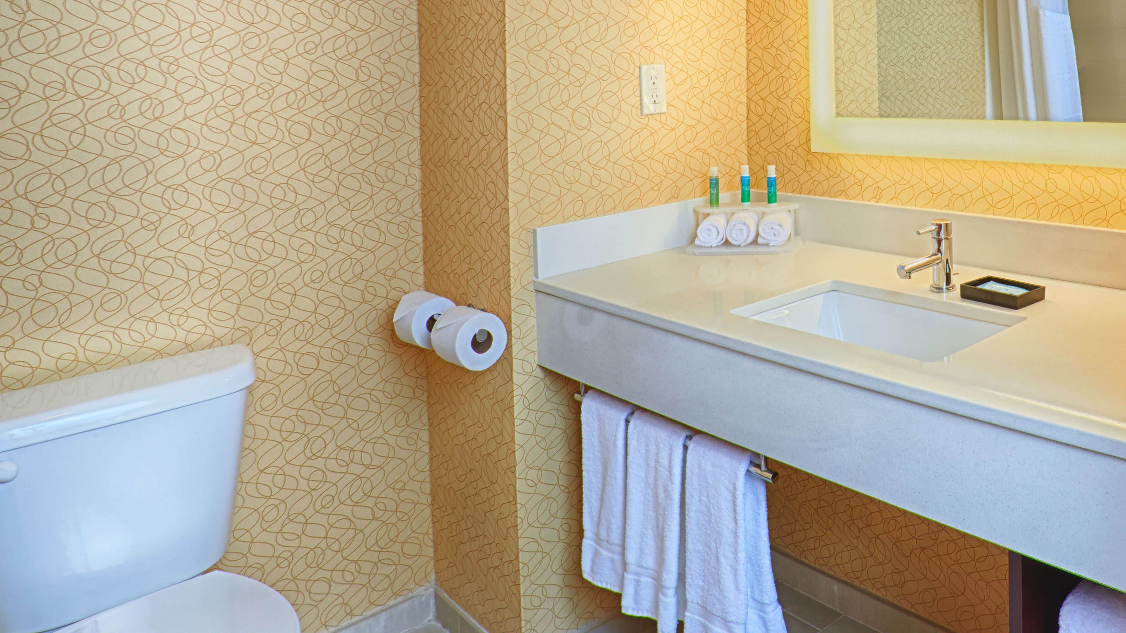 A bathroom with a toilet, a roll of toilet paper, a sink with toiletries, and a towel hanging on the wall.