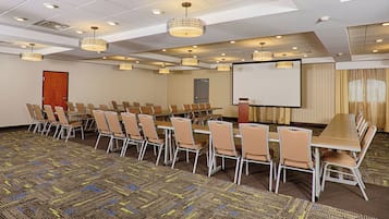 Sala de conferência com uma longa mesa, várias cadeiras, uma tela de projetor e luminárias no teto.
