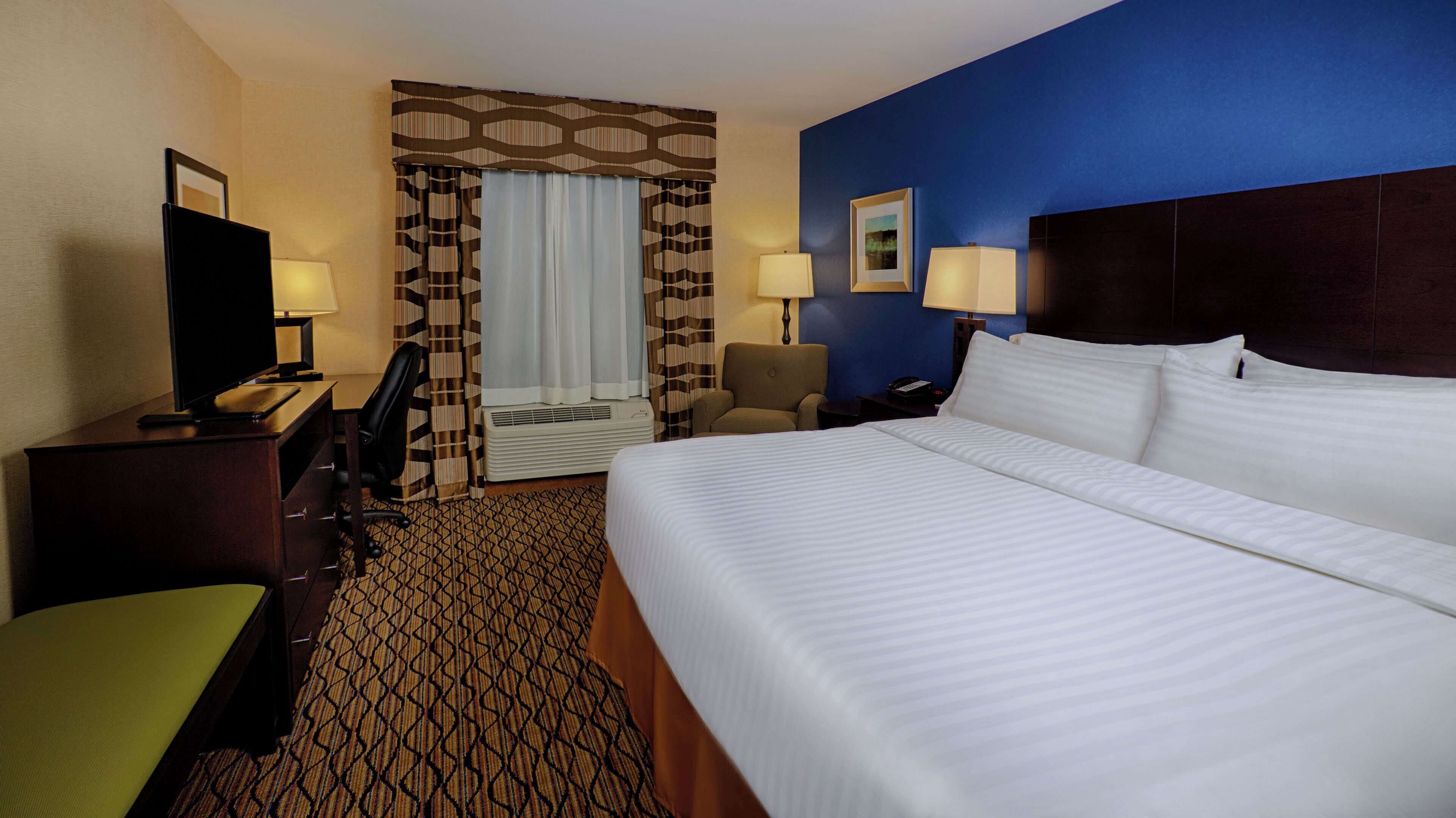Habitación de hotel con una cama grande, un escritorio con computadora, una silla, una lámpara de escritorio, un teléfono y una ventana con cortinas.