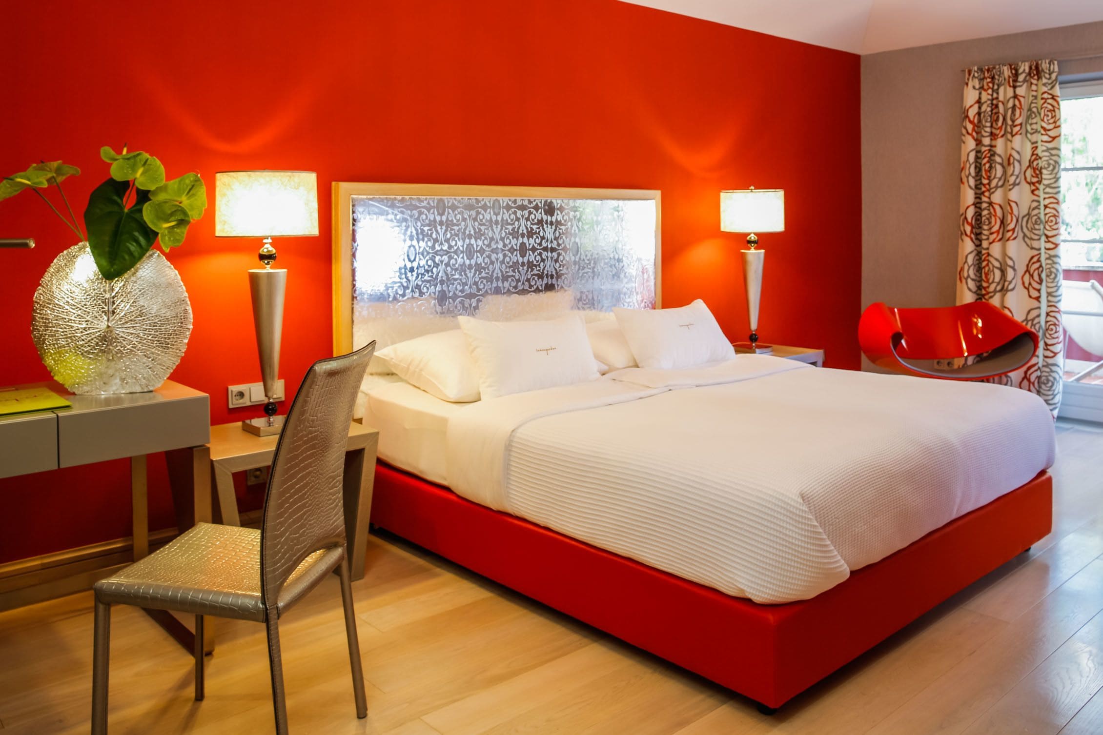 Una habitación de hotel moderna con una pared de acento roja, una cama grande con ropa de cama blanca, dos lámparas de noche, una silla roja y un escritorio con lámpara y un jarrón decorativo.