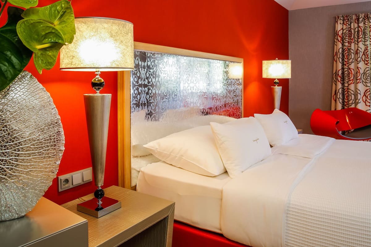 Hotellihuone, jossa on sänky, kaksi yöpöytälamppua, punainen tuoli ja koristeellinen kasvi.