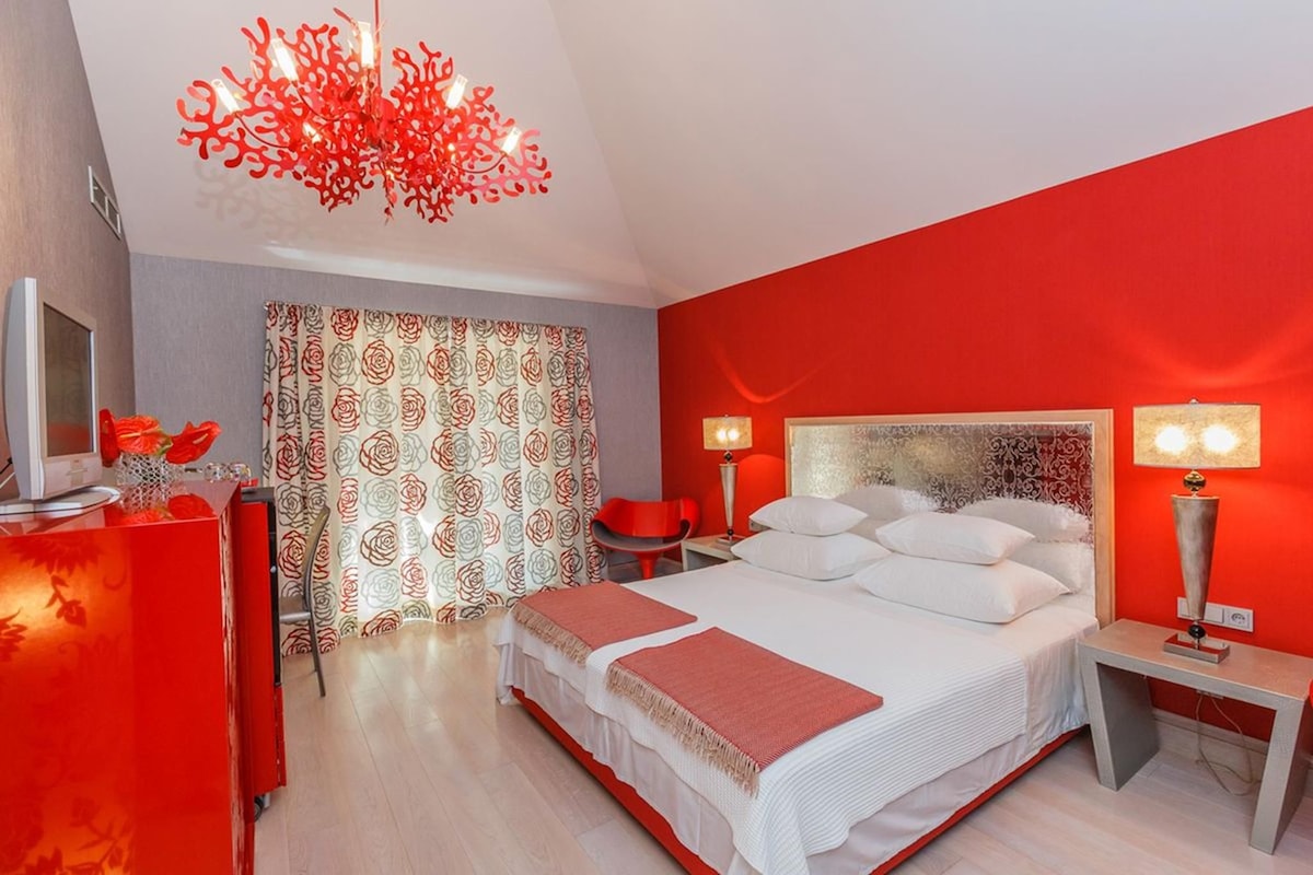 A modern hotel room with a large bed, a desk, a television, and a red accent wall.