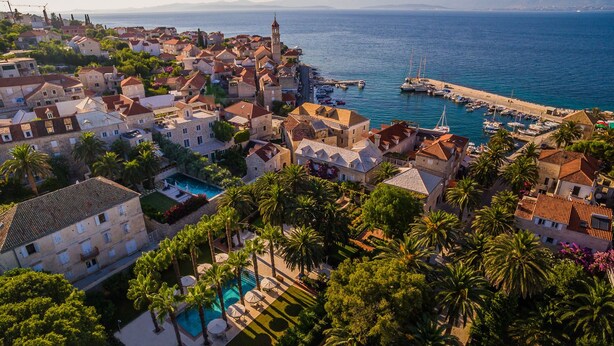 Une ville côtière dotée de bâtiments historiques, d’un port de plaisance et d’une piscine entourée de palmiers.