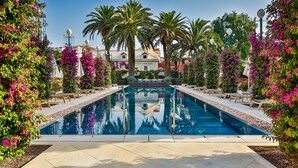 Une piscine entourée de palmiers et de fleurs roses, avec des chaises longues et un ciel bleu azur.