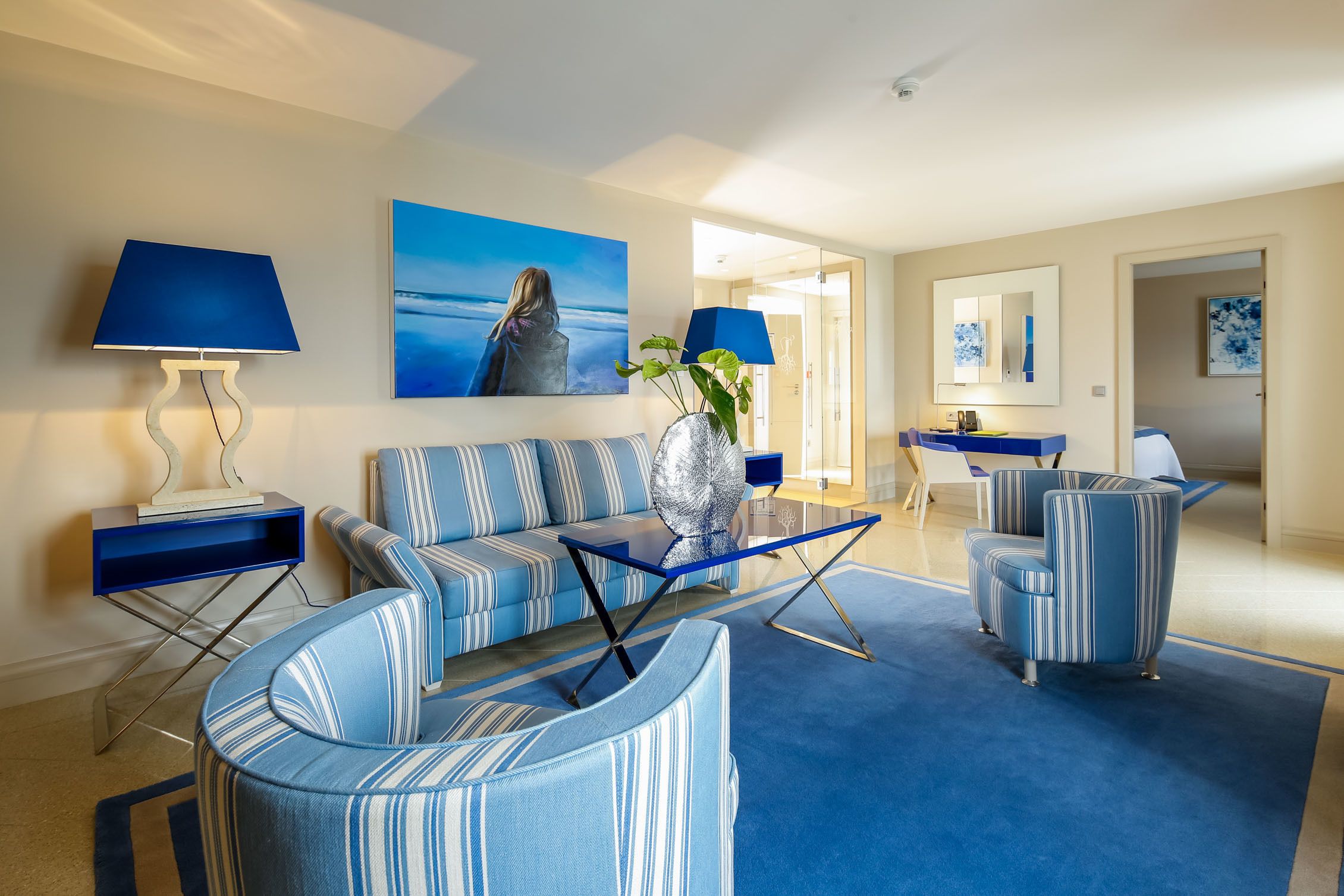 Un salón moderno con un sofá azul, sillones a rayas, una mesa de centro y un cuadro de una mujer junto al mar.