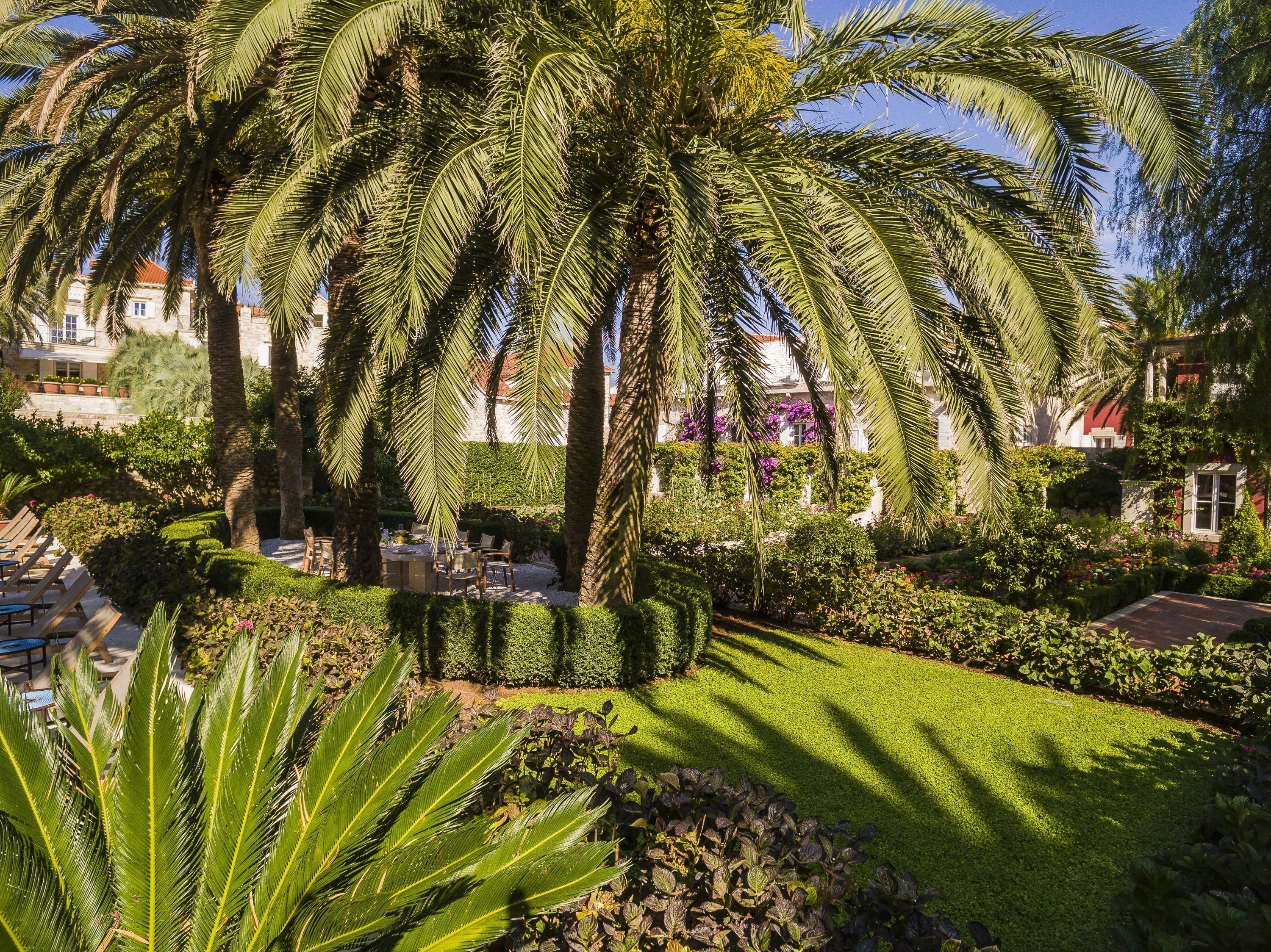 Un giardino curato con palme, un prato ben tagliato e un'area pranzo con tavoli e sedie.