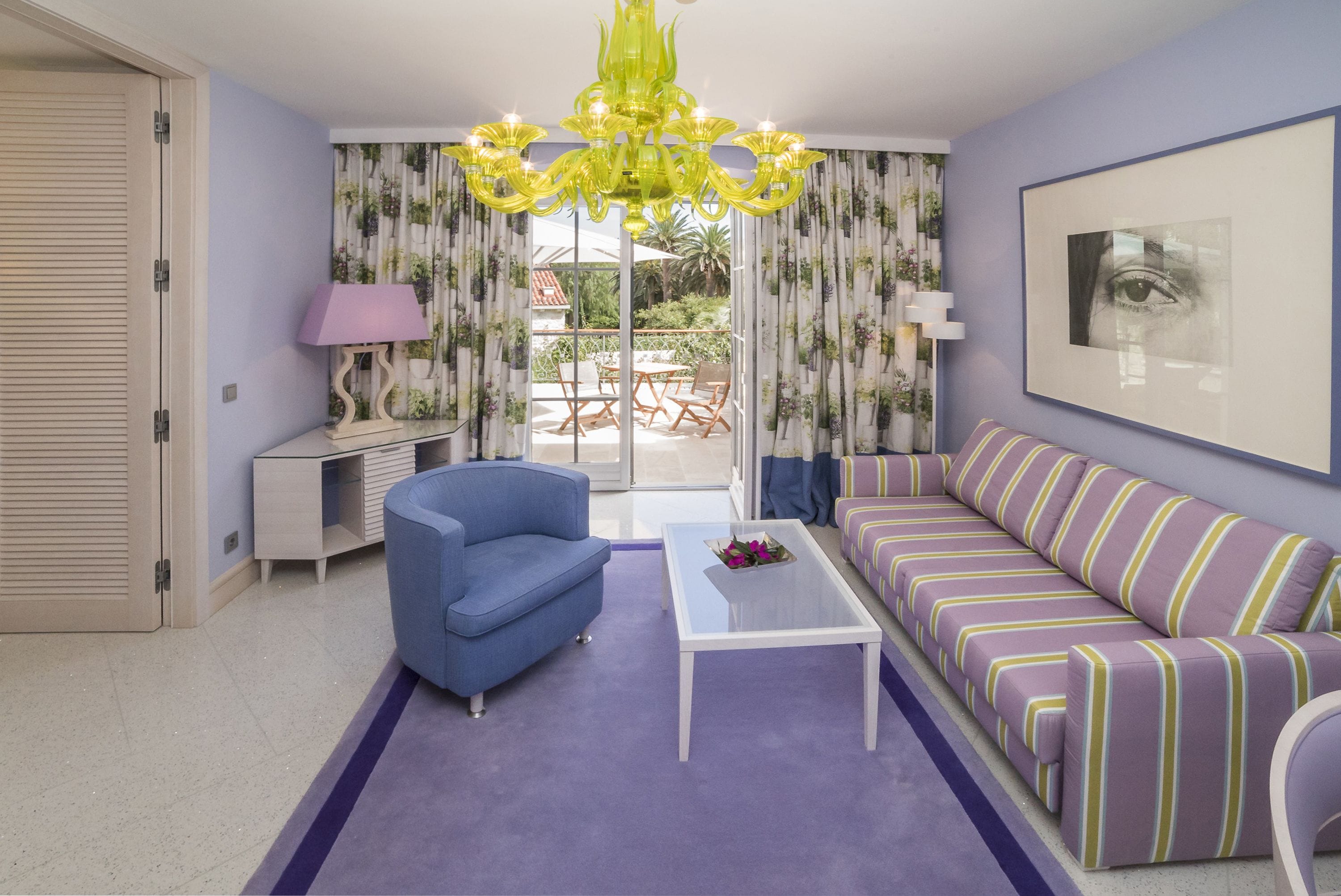 A living room with a blue armchair, a striped sofa, a glass coffee table, and a yellow chandelier.