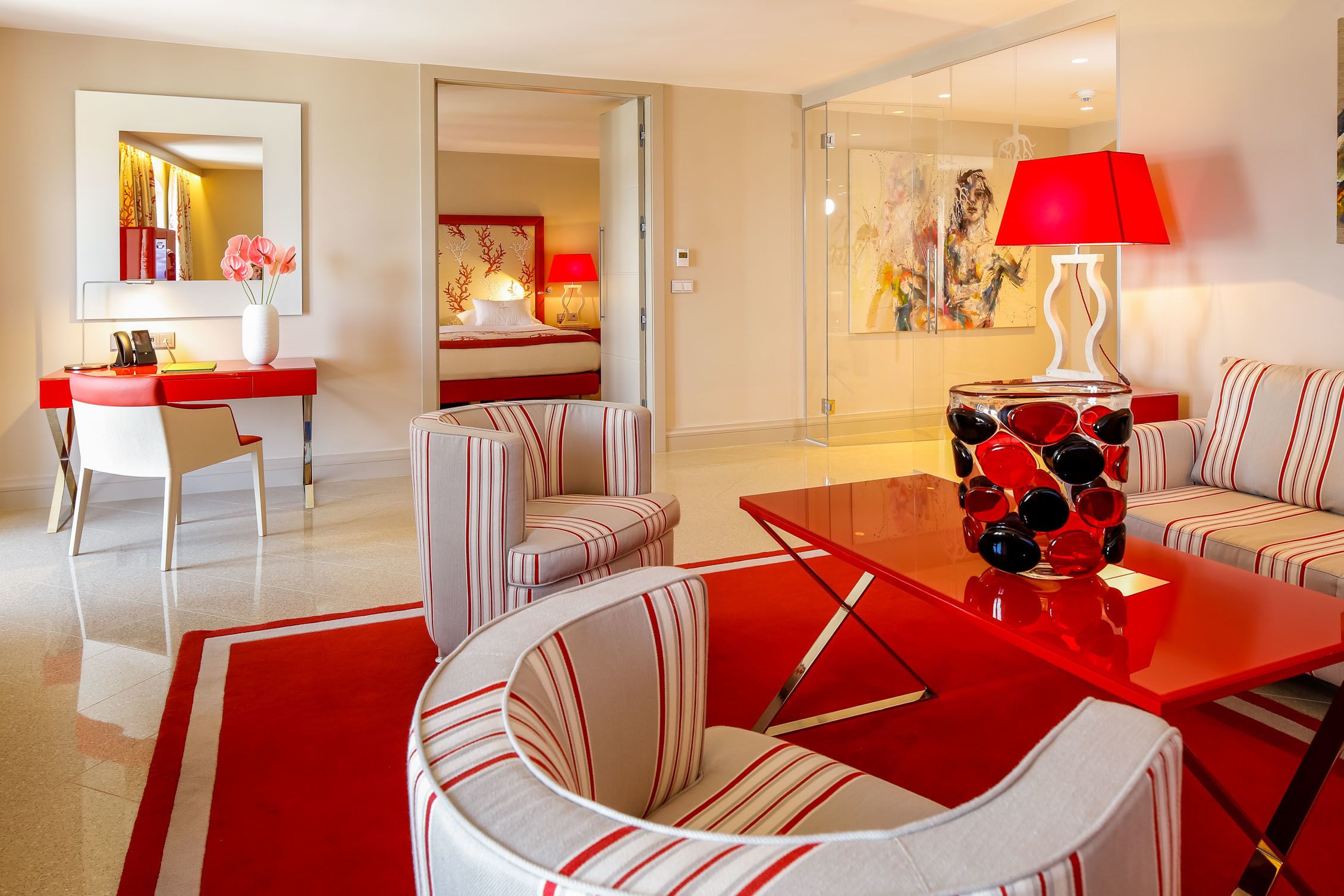 Una habitación de hotel moderna con una decoración en rojo y blanco, que incluye una ducha de vidrio, una zona de comedor con sillas a rayas y un escritorio con un teléfono y un jarrón de flores.