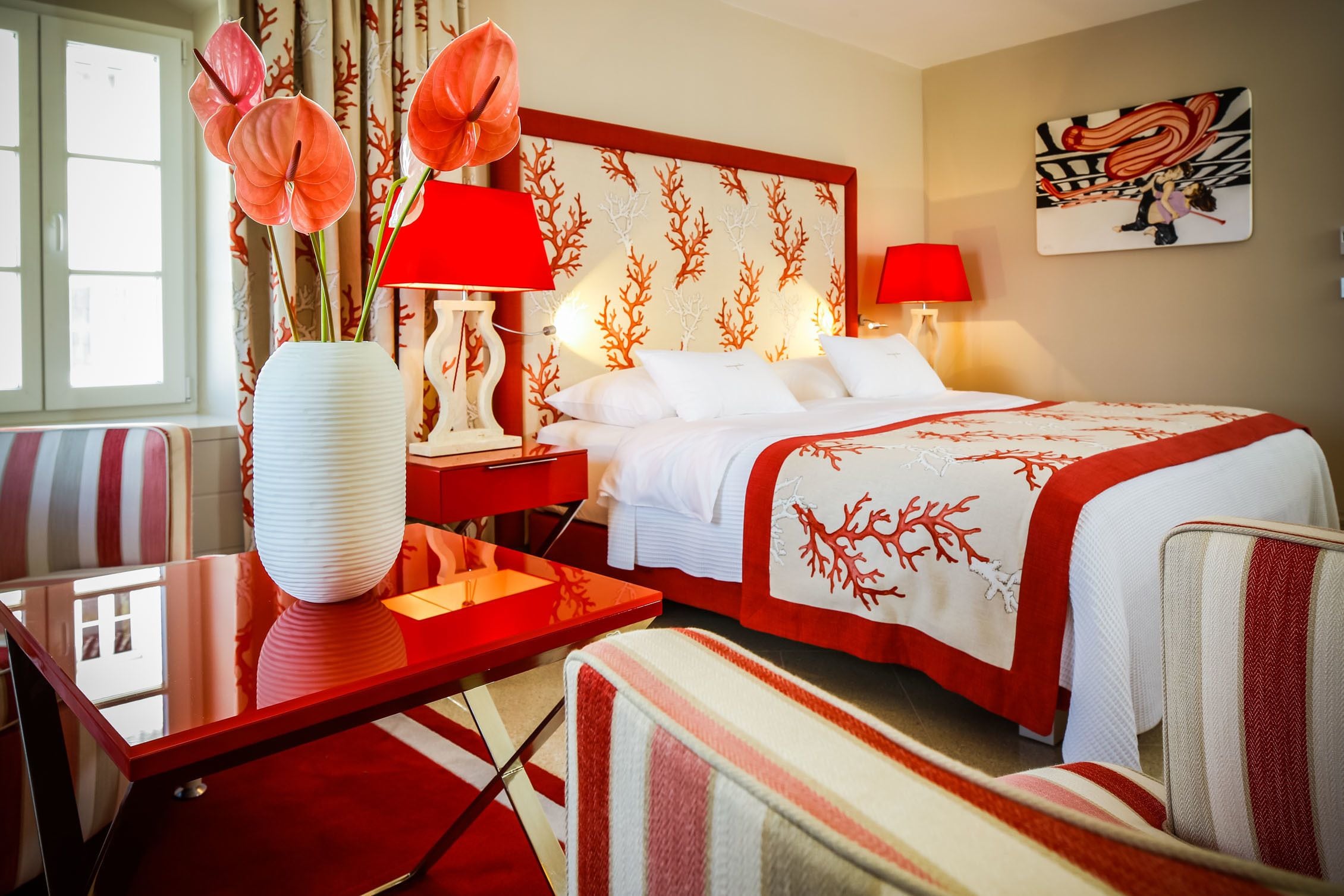 A hotel room with a bed, a desk, a chair, a vase with flowers, and a painting on the wall.