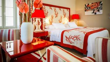 A hotel room with a bed, a desk, a chair, a vase with flowers, and a painting on the wall.