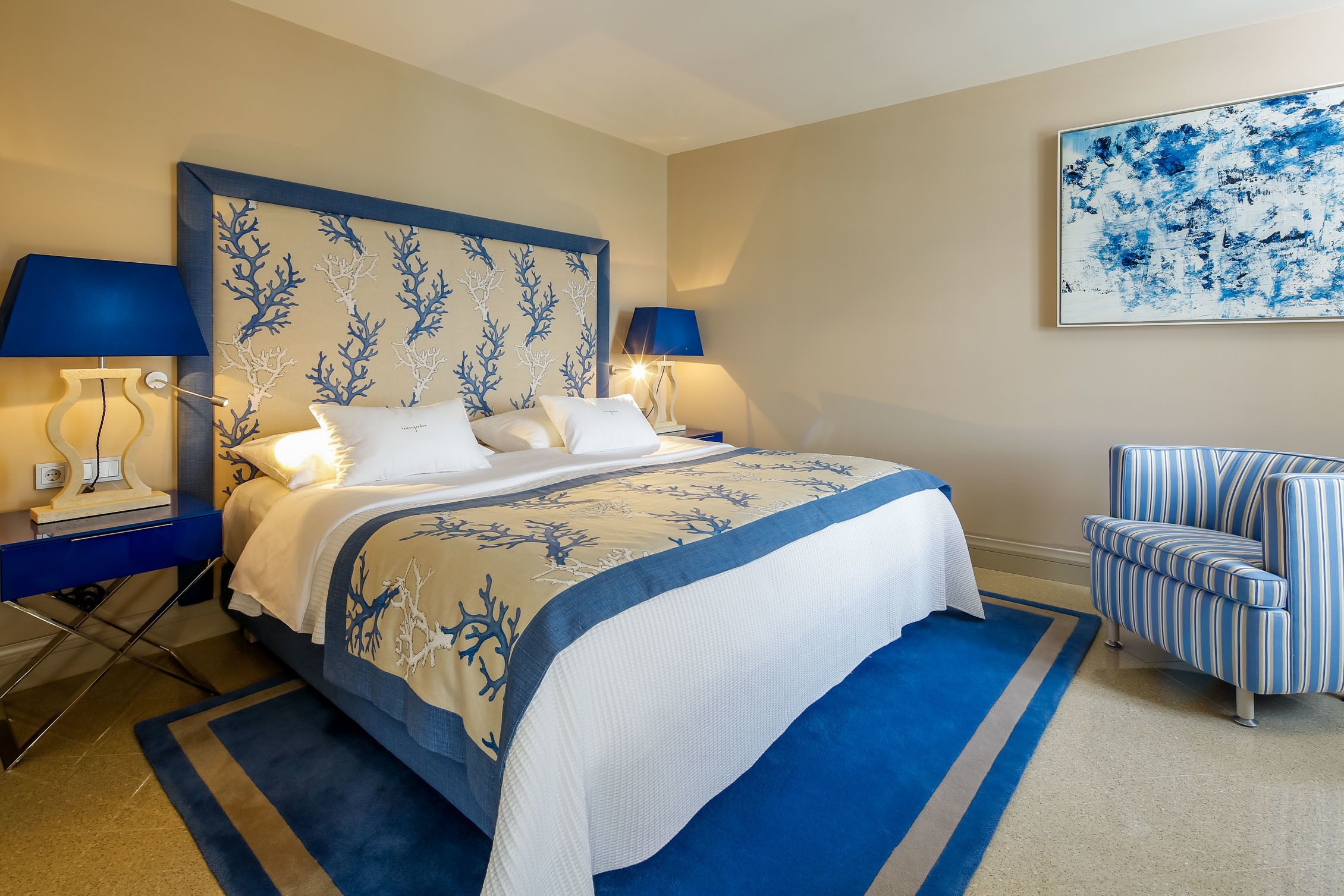 A neatly arranged hotel room with a large bed, two bedside tables with lamps, a striped armchair, and a large abstract painting on the wall.