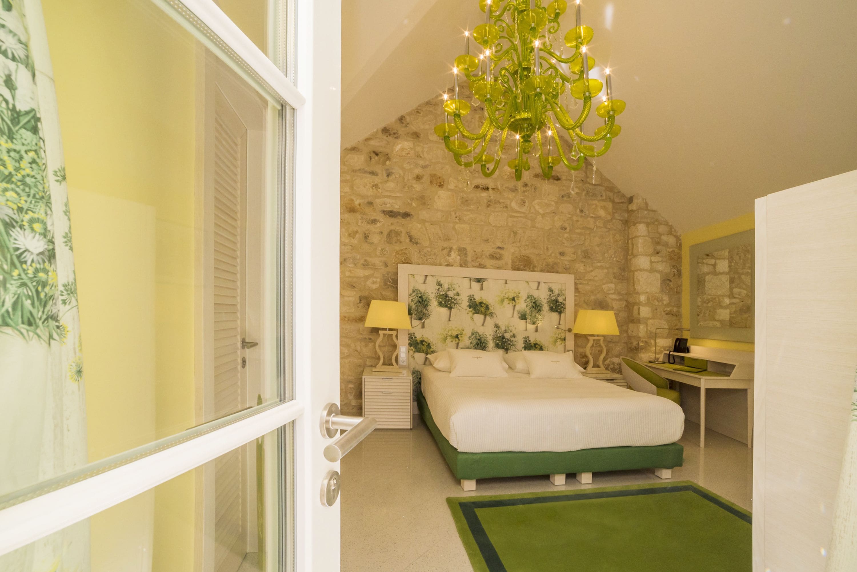 A bedroom with a stone wall, a bed with green bedding, a chandelier, and a desk with a lamp.