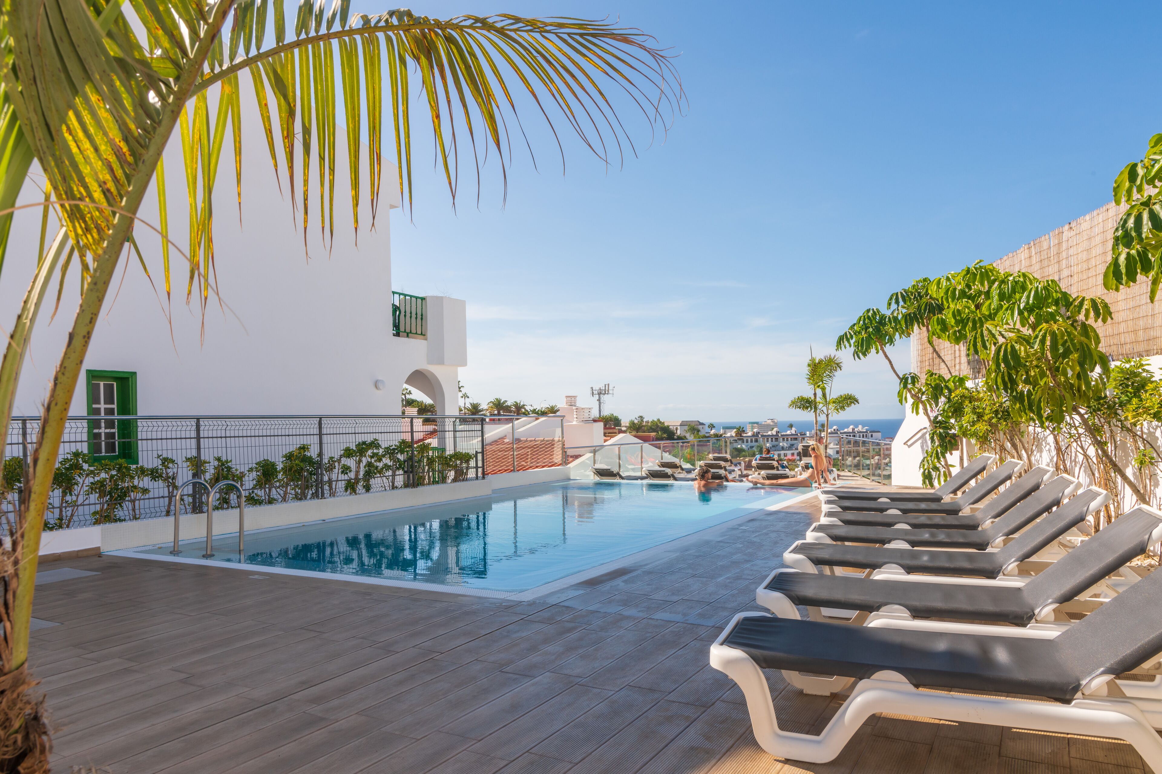 A rooftop pool area with lounge chairs, a clear view of the ocean, and a white building with green shutters.