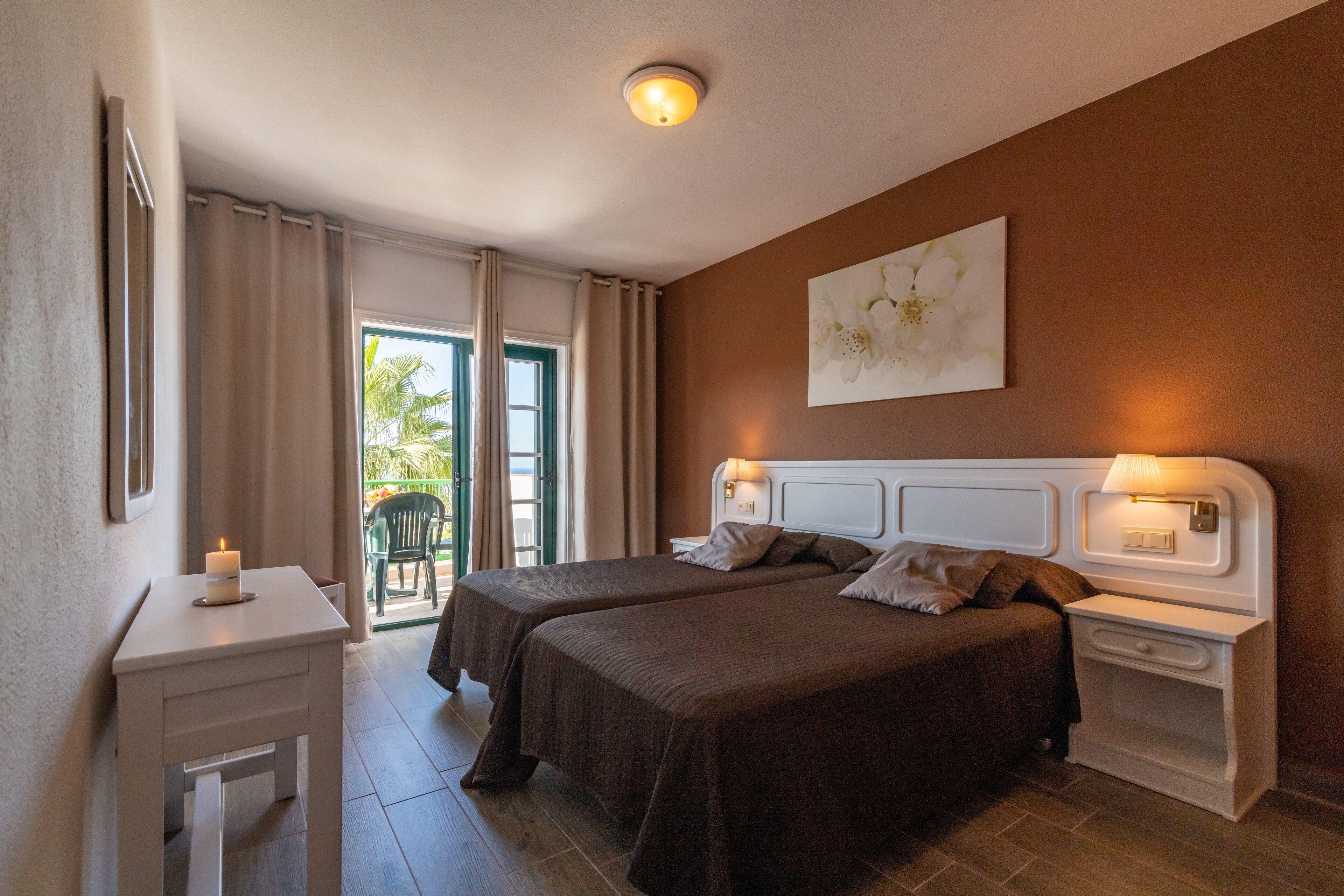 A hotel room with two beds, a small table with a candle, a nightstand with a lamp, and a painting on the wall.