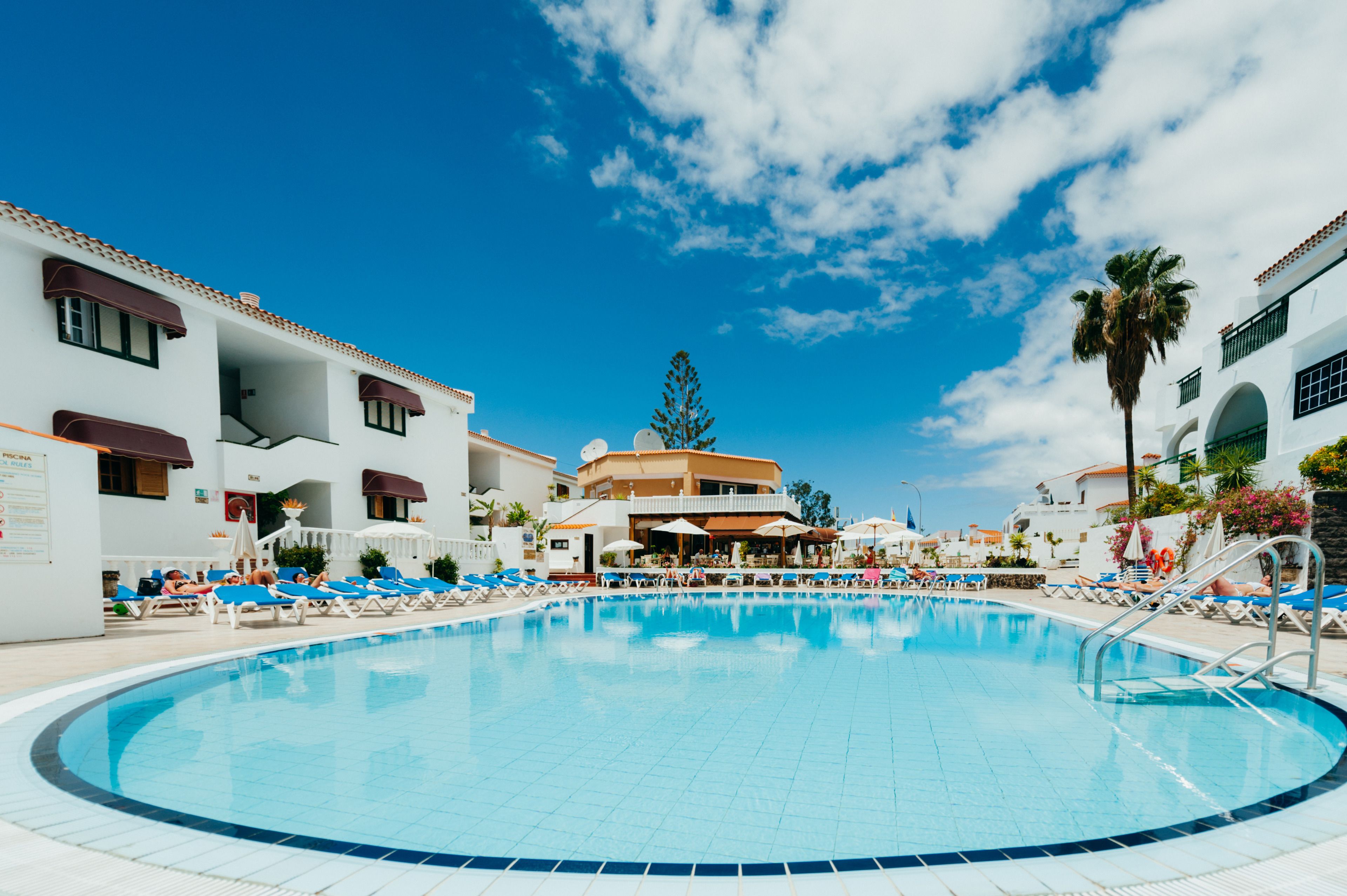 Una piscina con sillas de descanso y sombrillas, rodeada de edificios con balcones y ventanas.