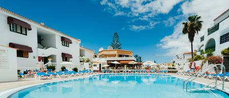 Una piscina con sillas de descanso y sombrillas, rodeada de edificios con balcones y ventanas.