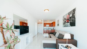 Ein modernes Wohnzimmer mit einer Couch, einem Couchtisch und einem Fernseher. Im Hintergrund ist eine Küche mit Schränken und einer Arbeitsplatte zu sehen.