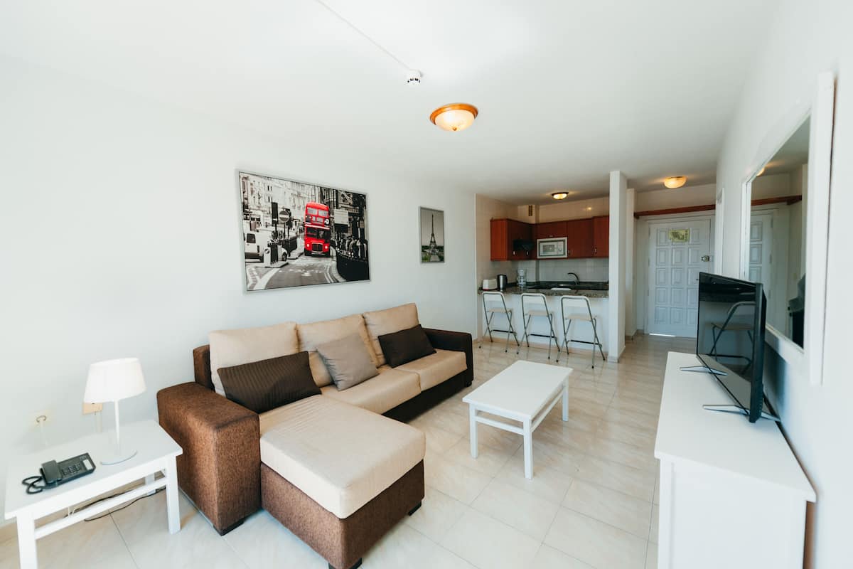 Un salon moderne avec un canapé, une table basse et un téléviseur. On aperçoit à l’arrière-plan un coin cuisine avec une table à manger et des chaises.