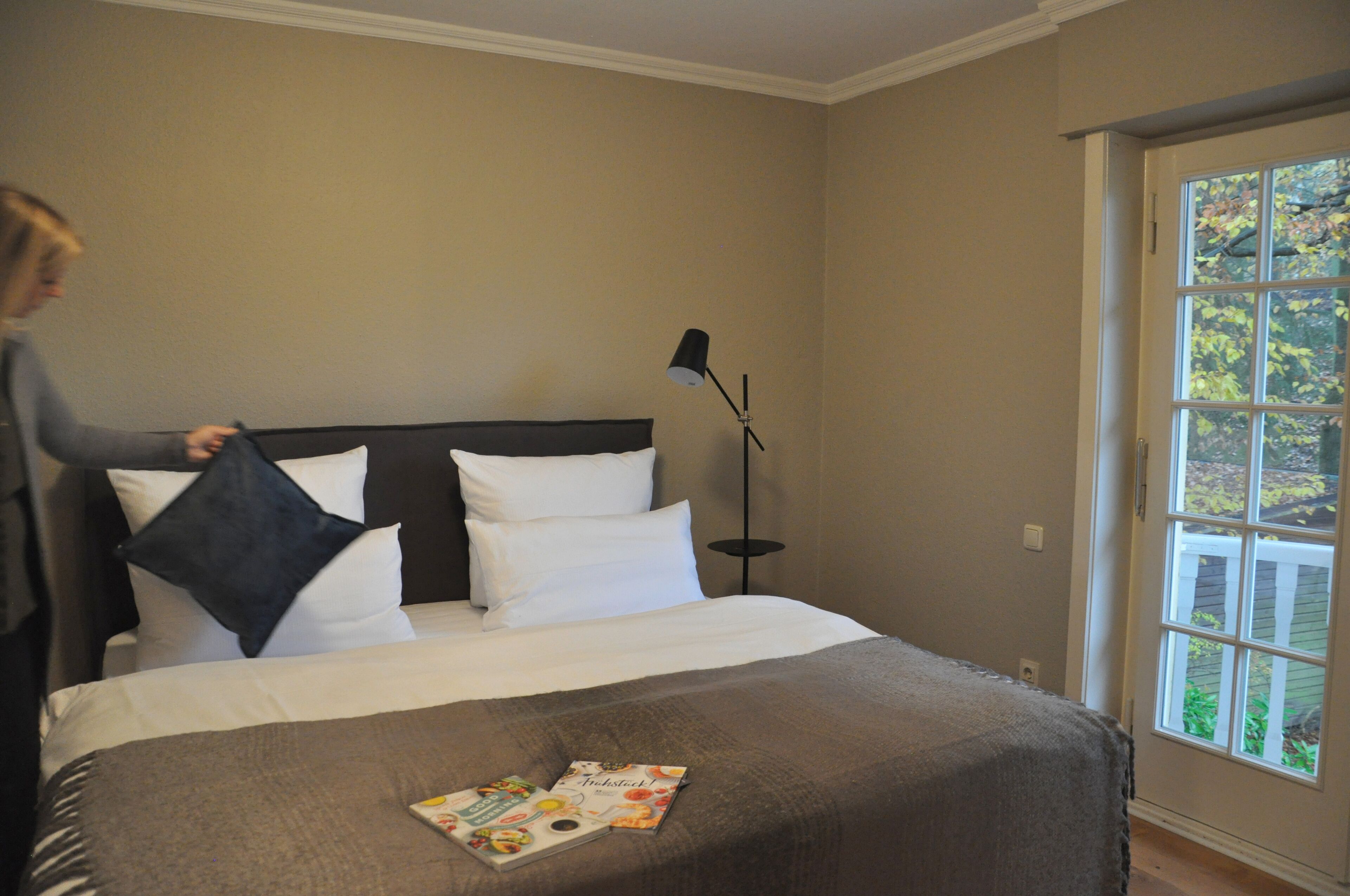 A neatly made bed with white and dark pillows, a magazine on the bed, and a floor lamp beside it.