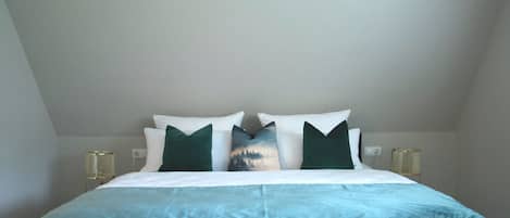 Una cama bien hecha con edredón azul y blanco, dos almohadas verdes y una almohada decorativa con una escena natural.