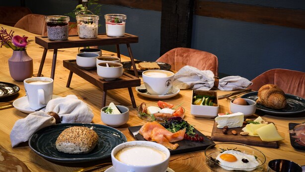 Une table de salle à manger en bois, sur laquelle sont disposés divers aliments pour le petit-déjeuner, notamment du pain, des œufs, du fromage et du saumon fumé.