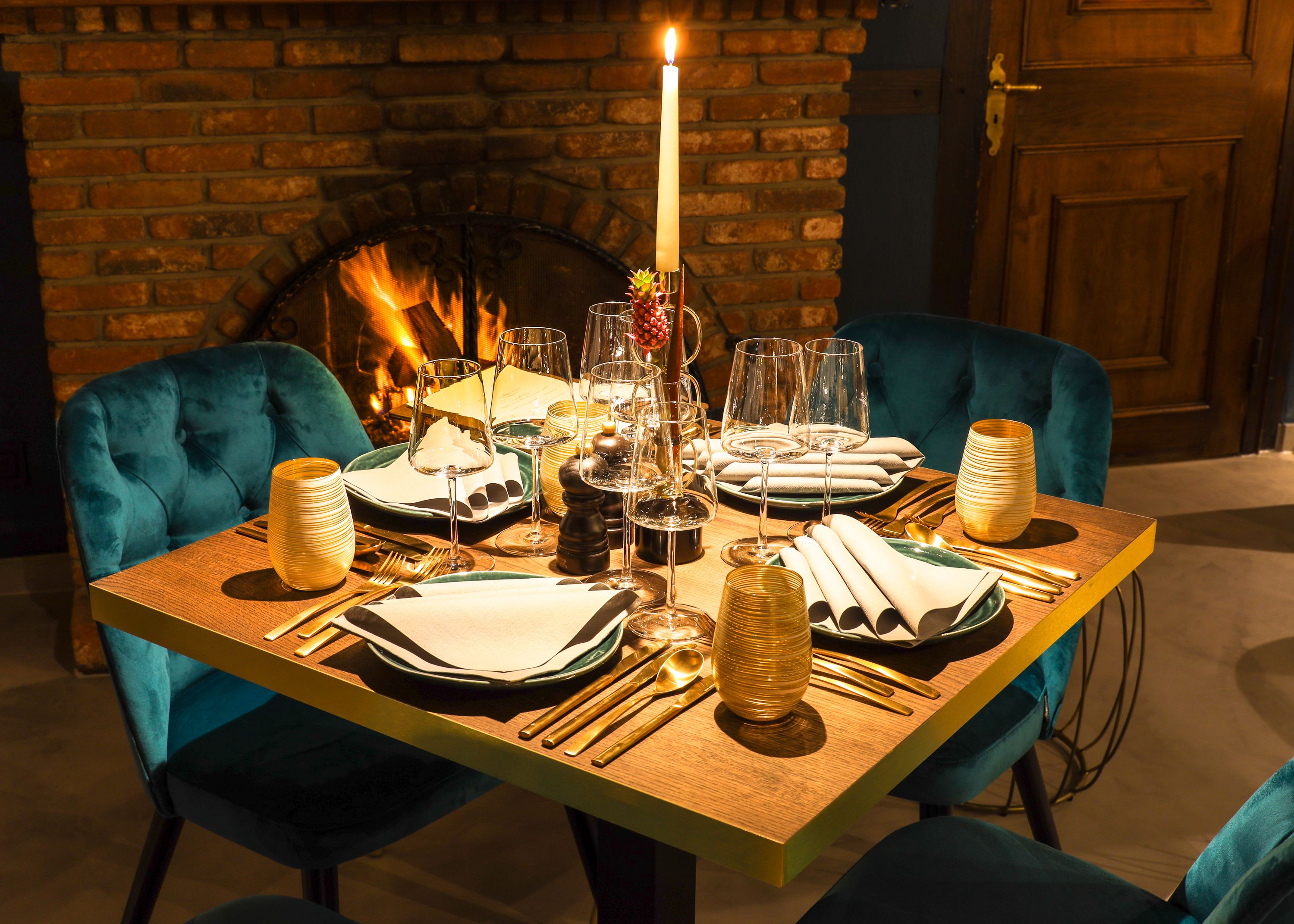 A dining table set for four with a candle centerpiece, surrounded by teal chairs, in front of a brick fireplace.