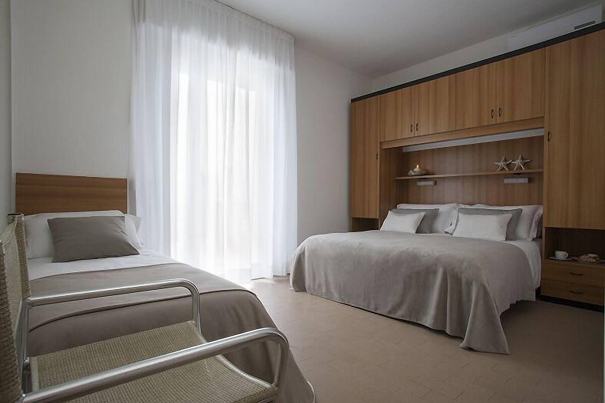 A hotel room with two beds, a wooden wardrobe, and a window with white curtains.