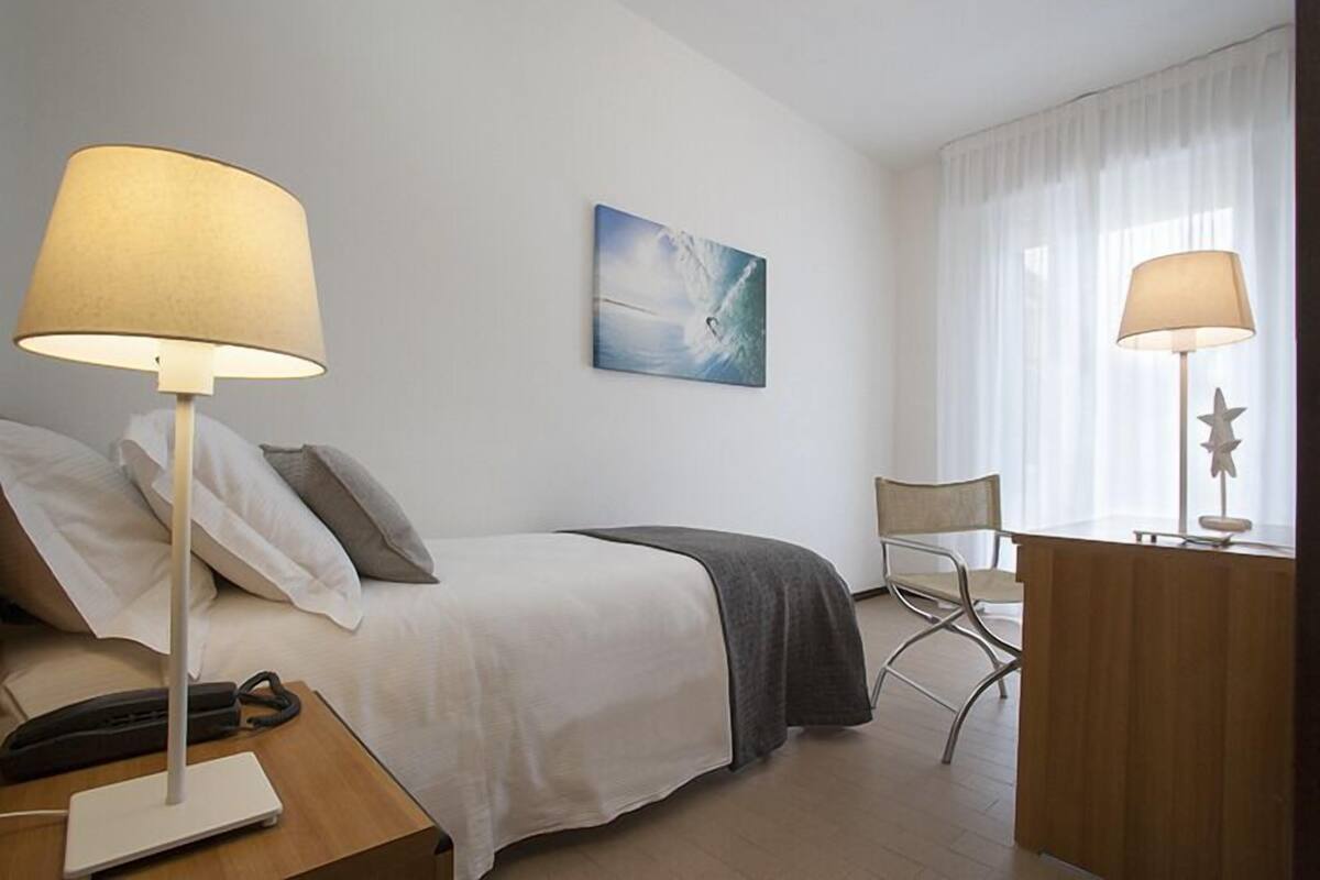 A hotel room with a bed, two bedside lamps, a desk with a lamp, a chair, and a painting on the wall.