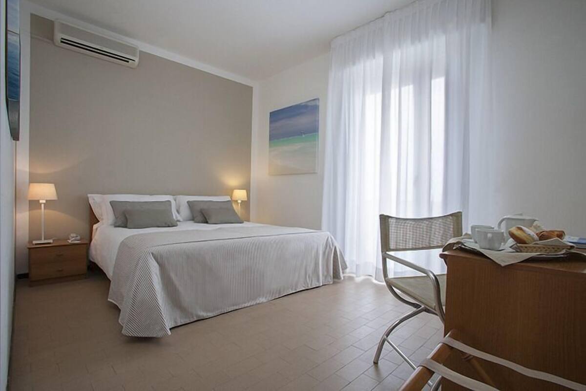 A hotel room with a bed, bedside lamps, a desk with a chair, and a window with curtains.